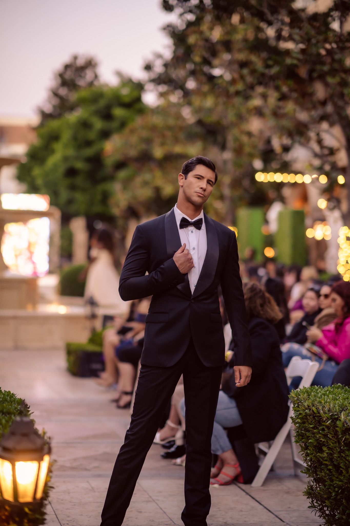 A group of groomsmen in black tuxedos pose playfully in a row at an outdoor evening wedding venue.