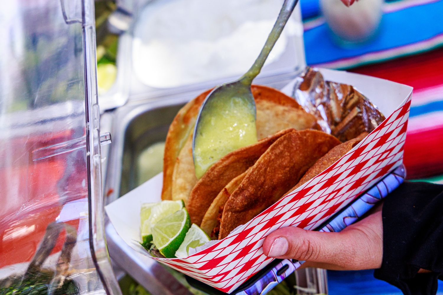 Hand holding a red checkered tray of crispy tacos with lime wedges and green salsa at a food stand.