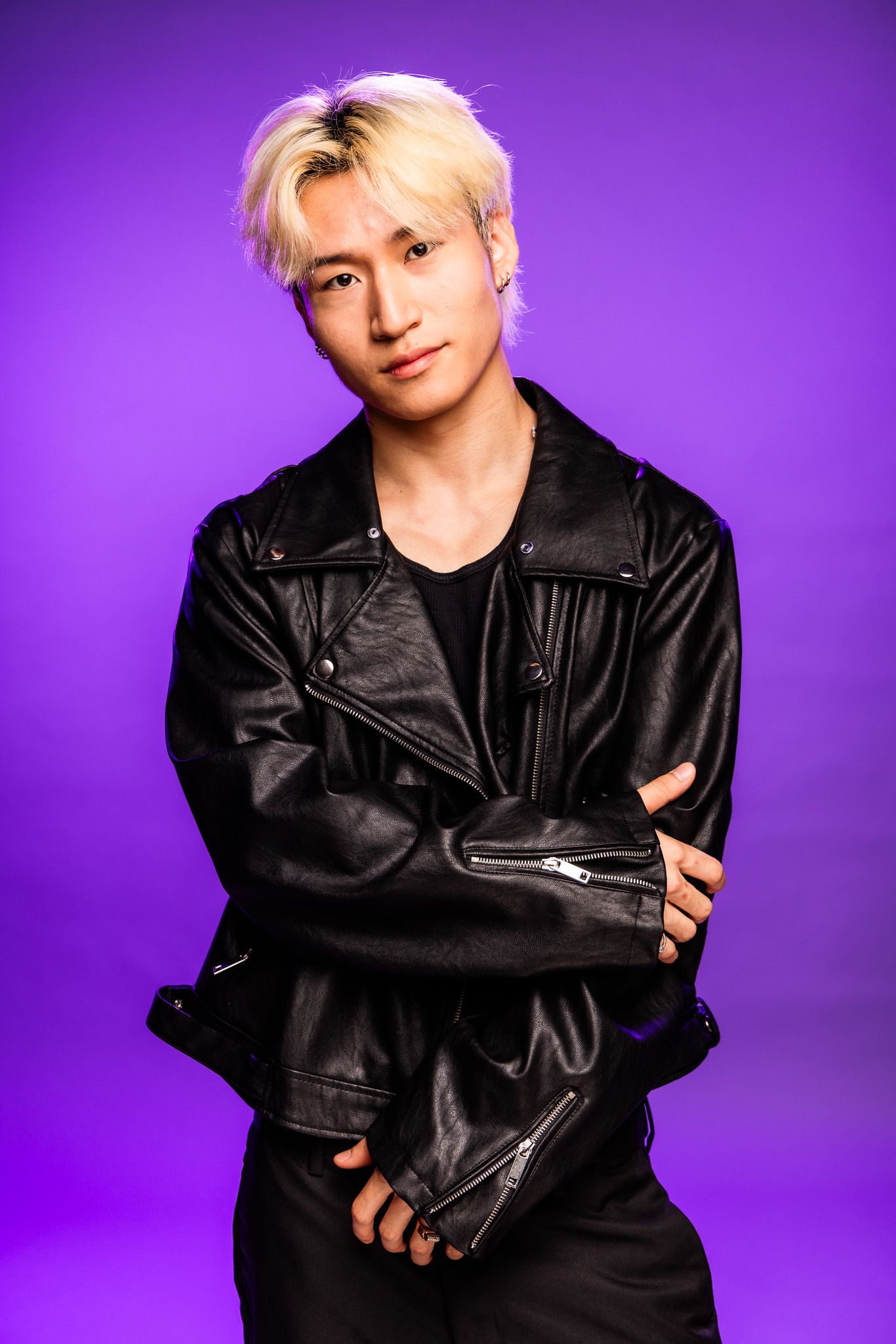 Young man with blonde hair wearing a black leather jacket, posing confidently against a vibrant purple background.