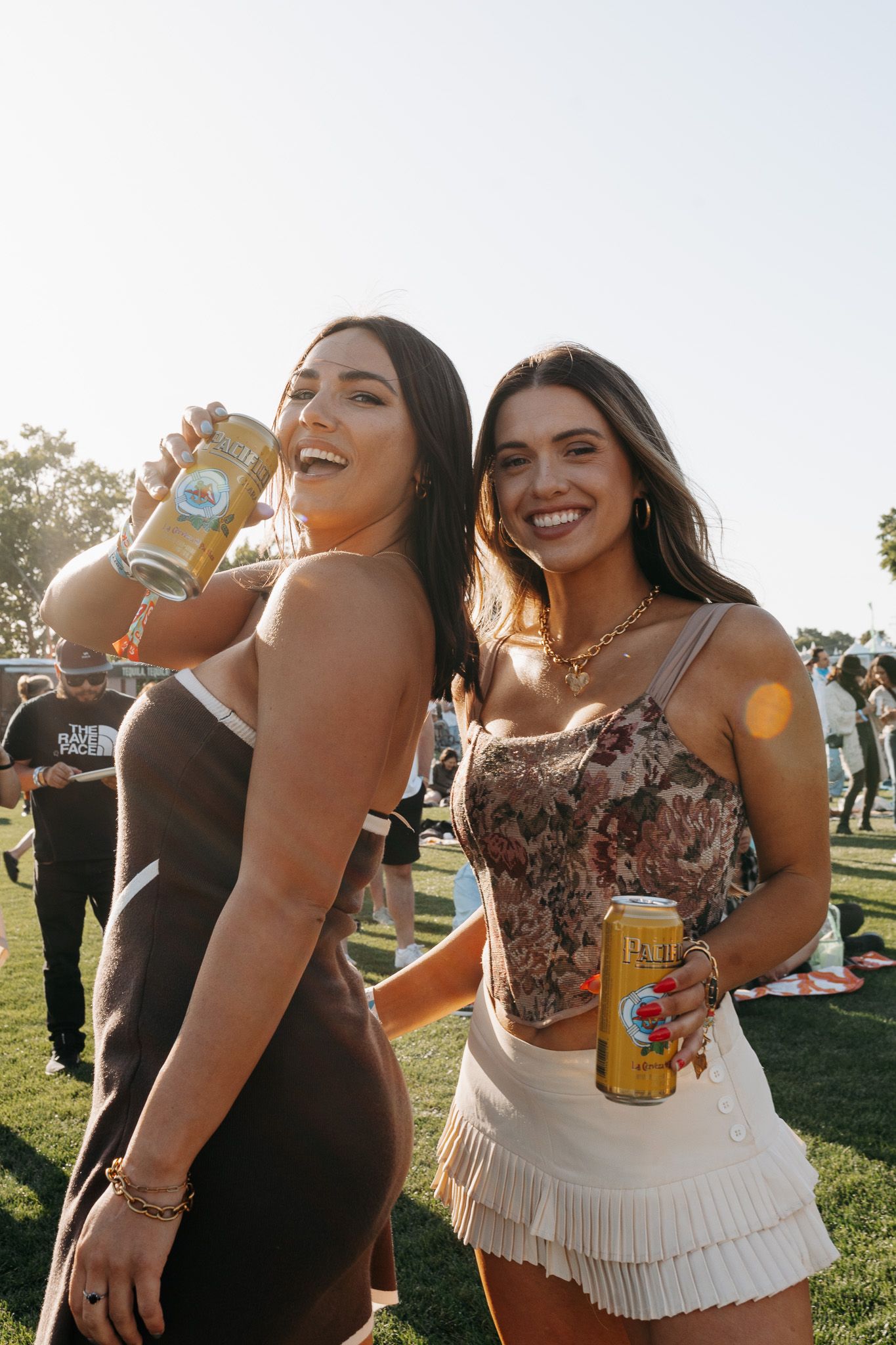 Two smiling women holding canned drinks at an outdoor festival on a sunny day.