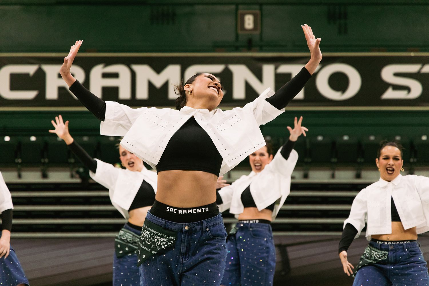 Dancers in white jackets and denim perform energetically at Sacramento State arena, with lead dancer striking a dramatic pose.