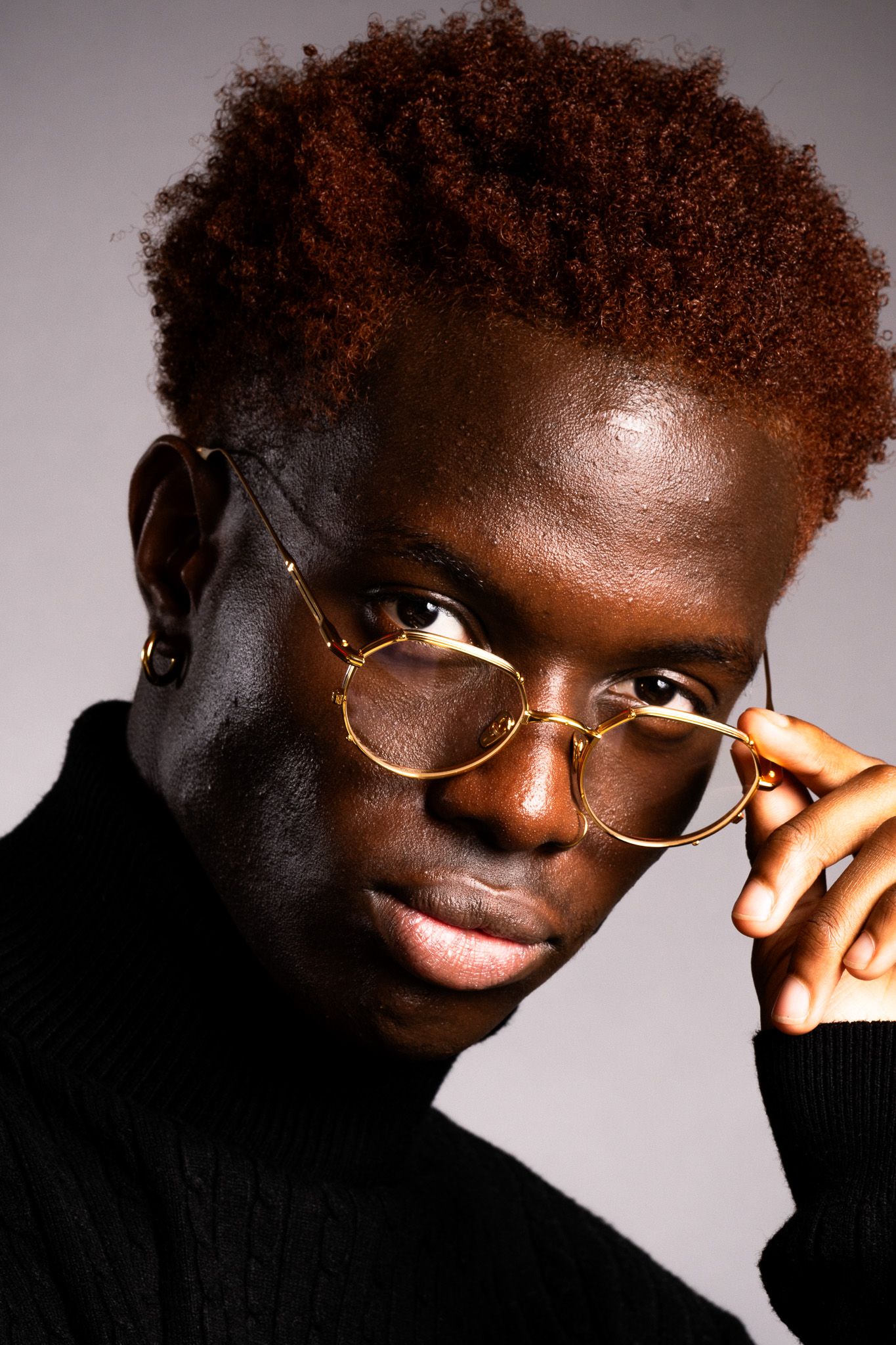 Young Black man with red afro hair wearing gold round glasses and black turtleneck, studio portrait.