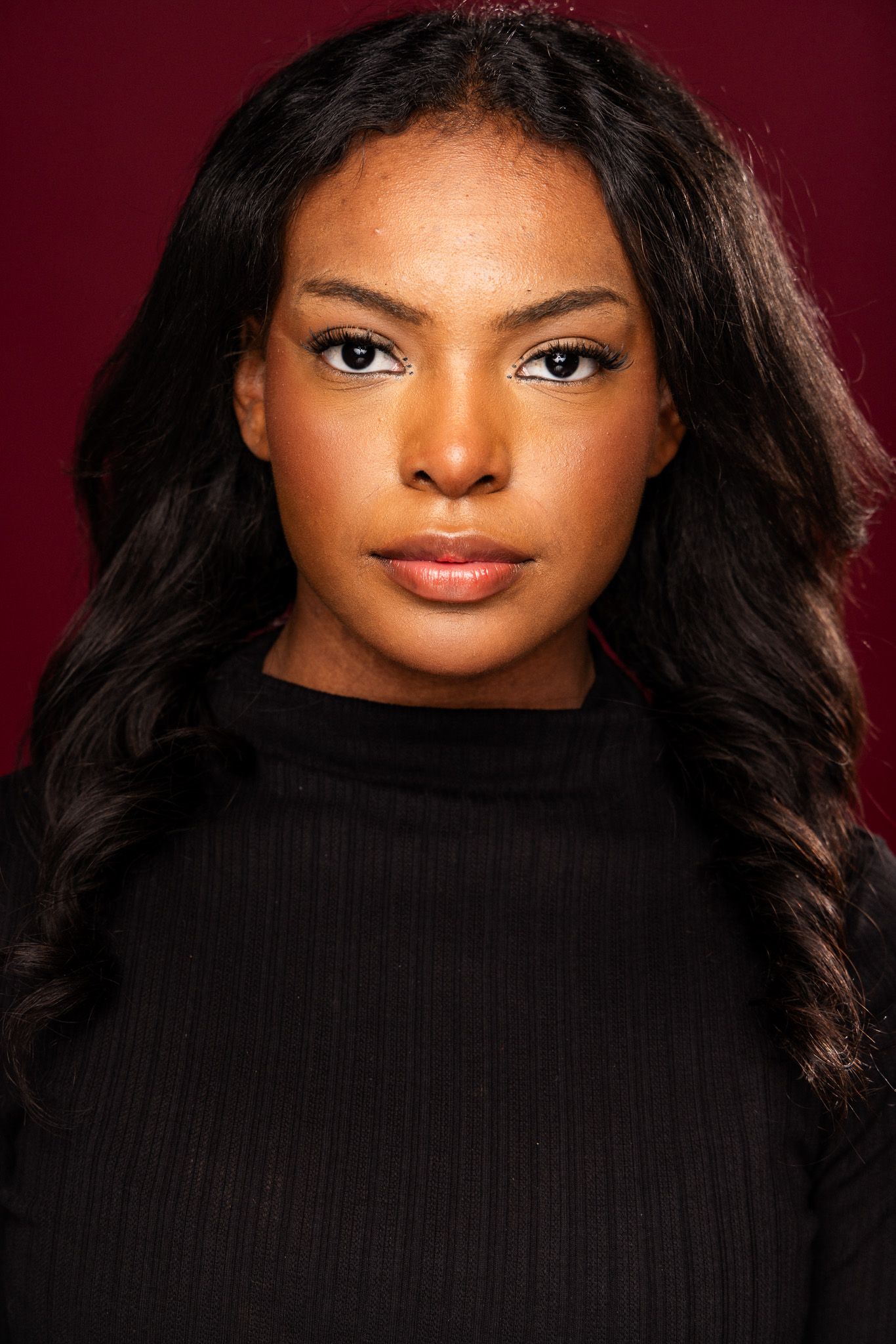 Portrait of a woman with long dark hair wearing a black turtleneck against a dark burgundy background.