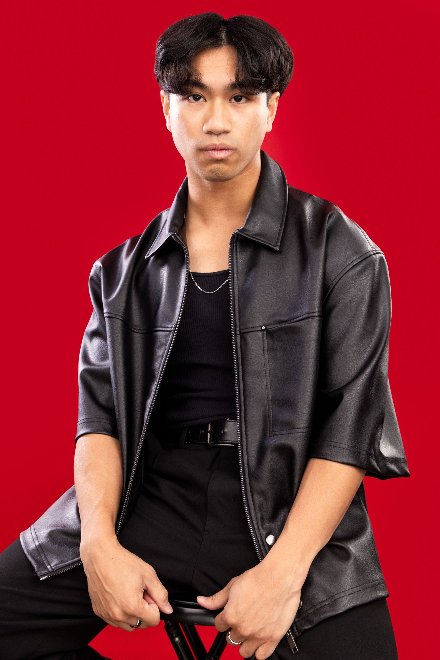 Young person in black leather jacket and t-shirt poses against vibrant red background in studio photoshoot.