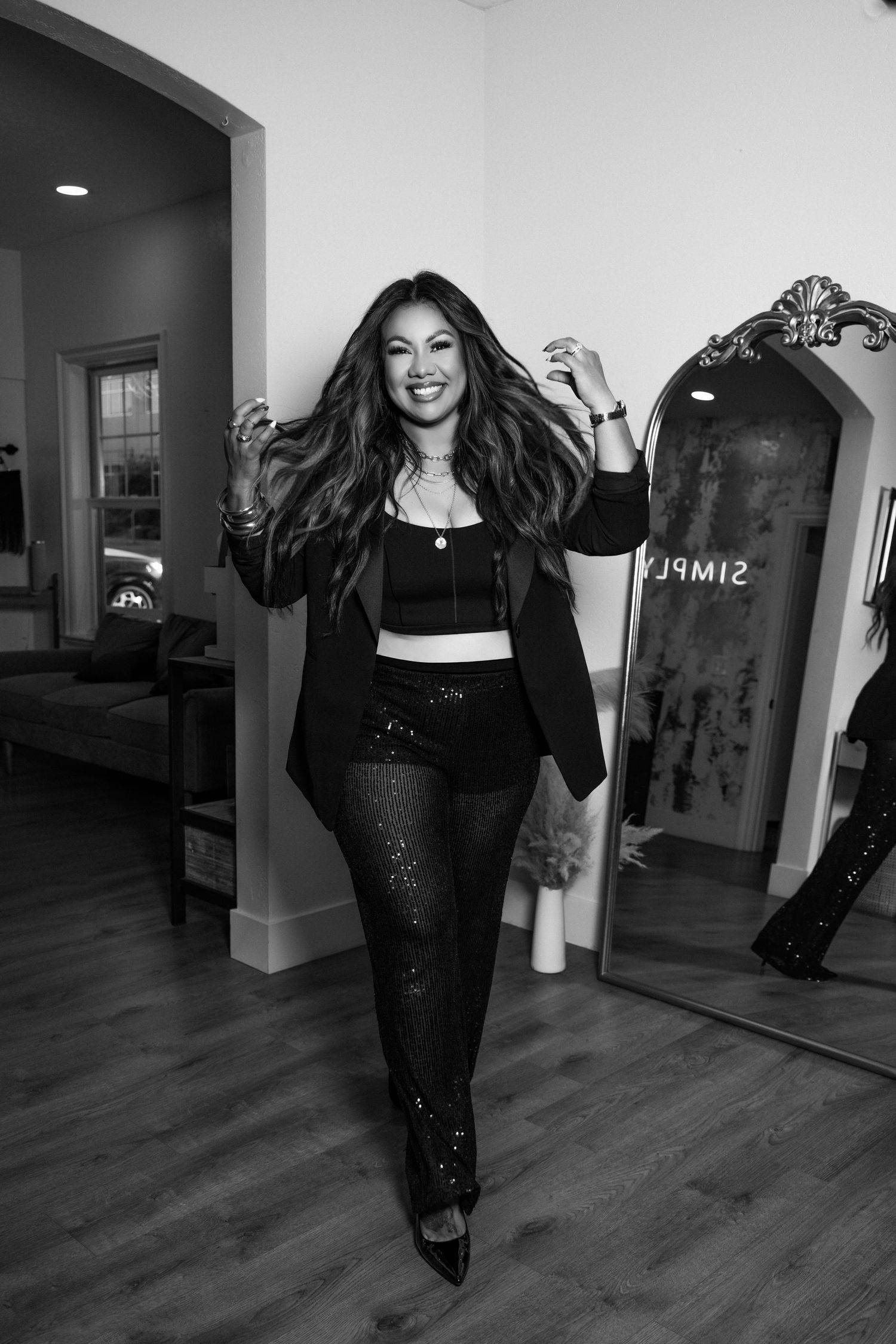 A person in a black blazer and sparkly pants poses energetically in a hallway with arched doorways and hardwood floors.