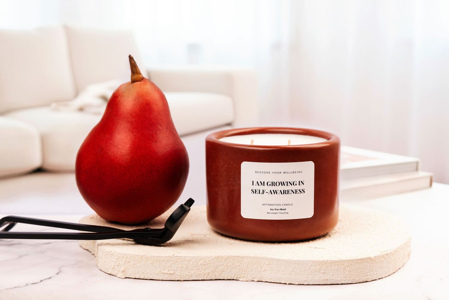 A minimalist composition featuring a red pear and a white candle on a light beige surface in soft natural lighting.