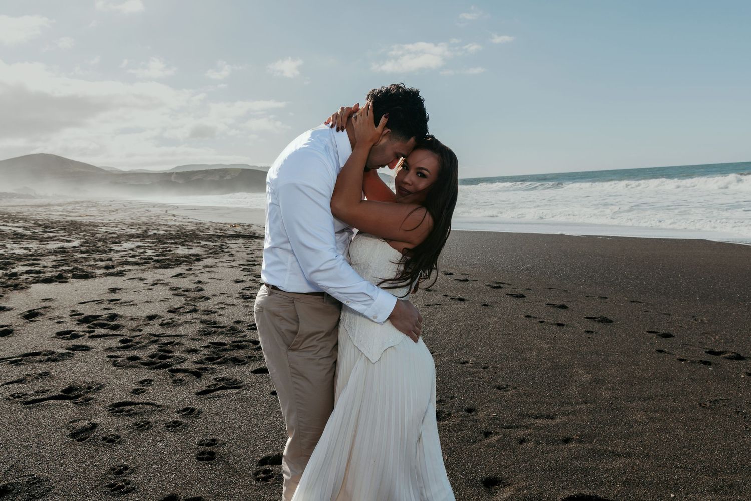 Romantic couple embraces on a serene beach with waves and mountains in the background during golden hour.