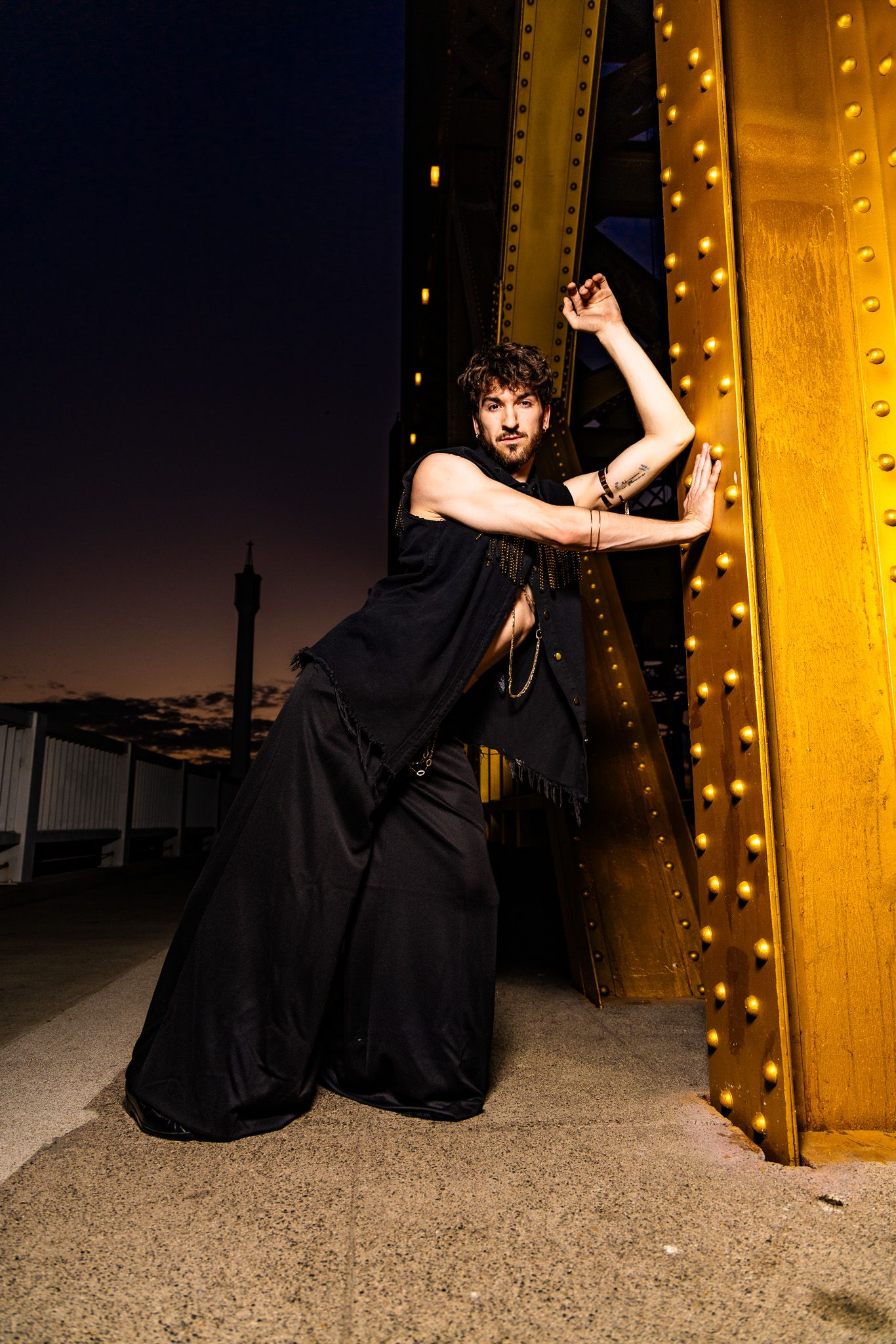 A person in black dress sits near an illuminated yellow wall in dramatic poses during an outdoor photoshoot.