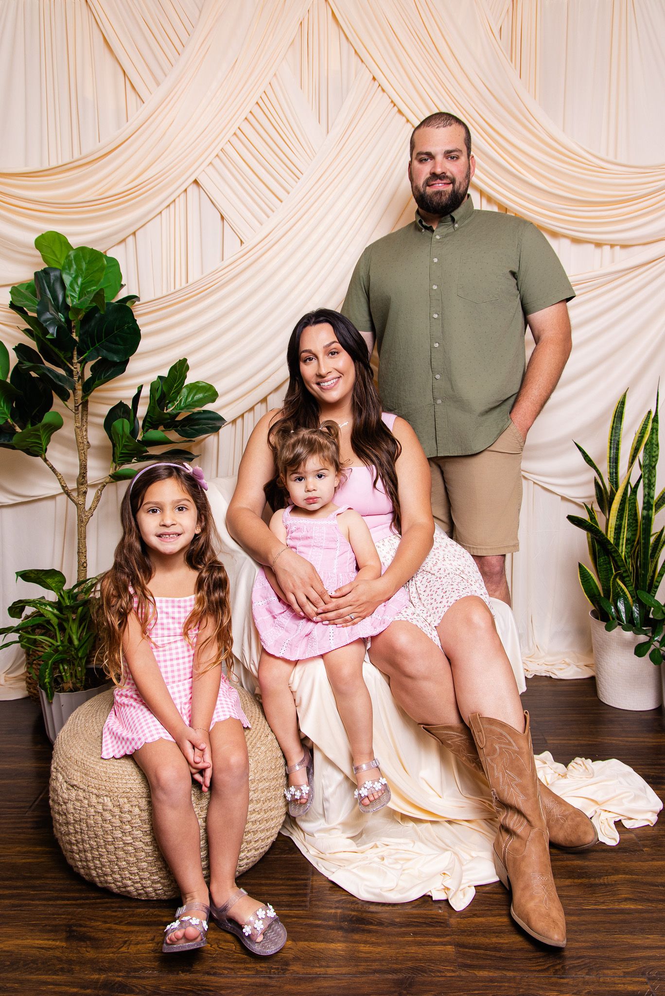 A happy family of four poses for a portrait with cream drape backdrop, green plants, featuring parents and two young girls in pink.