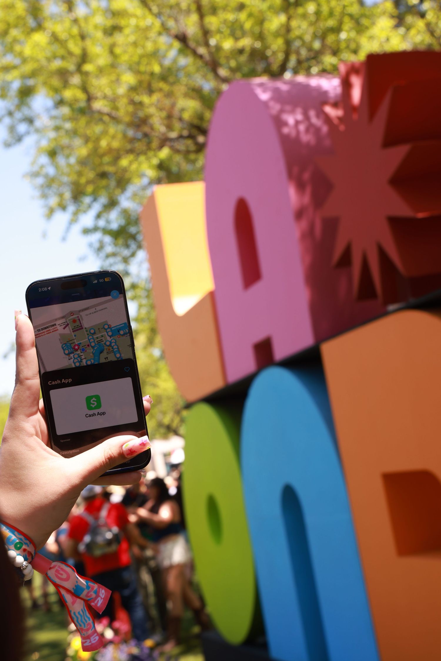Hands holding smartphones to photograph colorful outdoor letters against trees and blue sky in background.