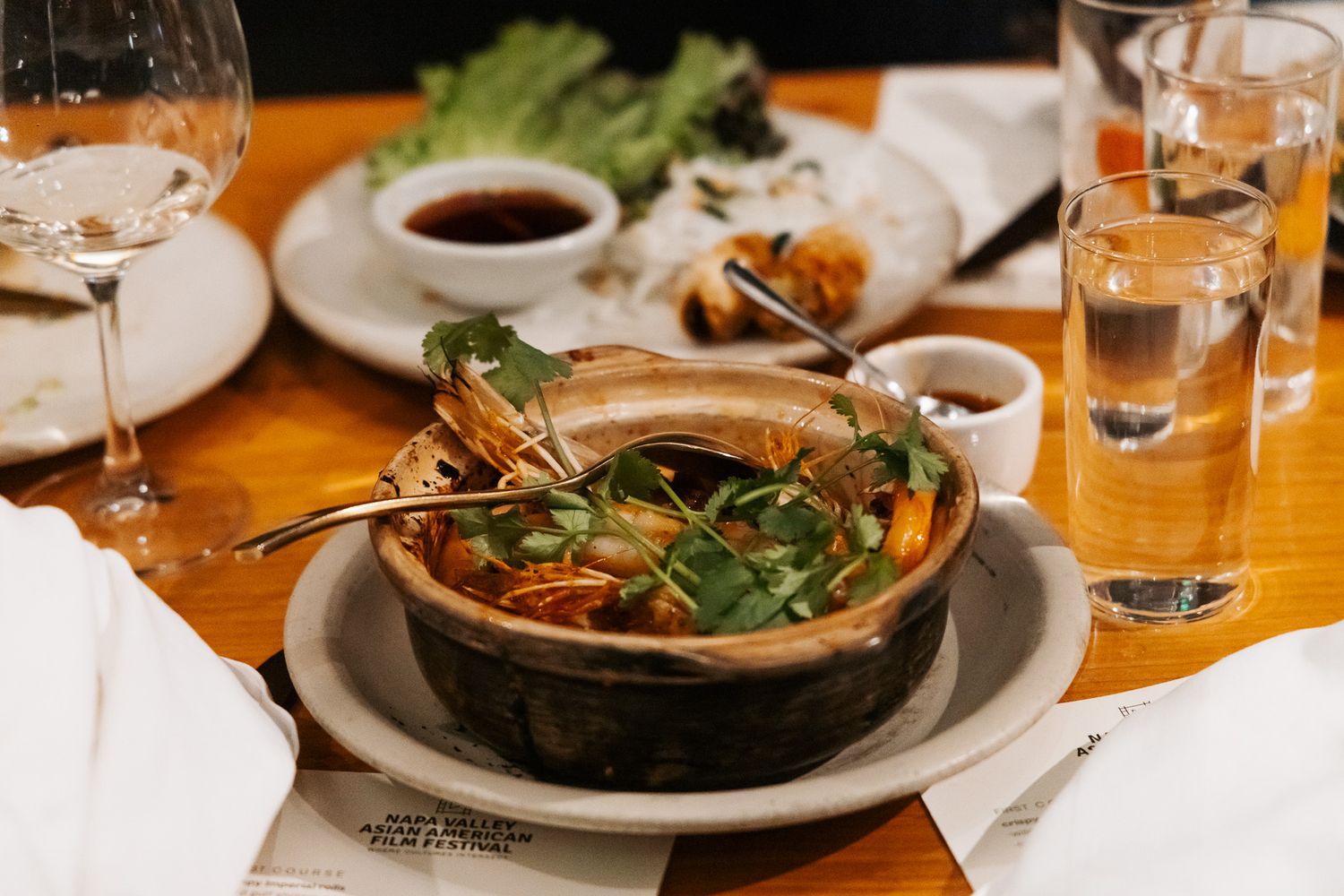 A steaming hot bowl of Asian soup garnished with fresh herbs served on a wooden table with wine glasses.