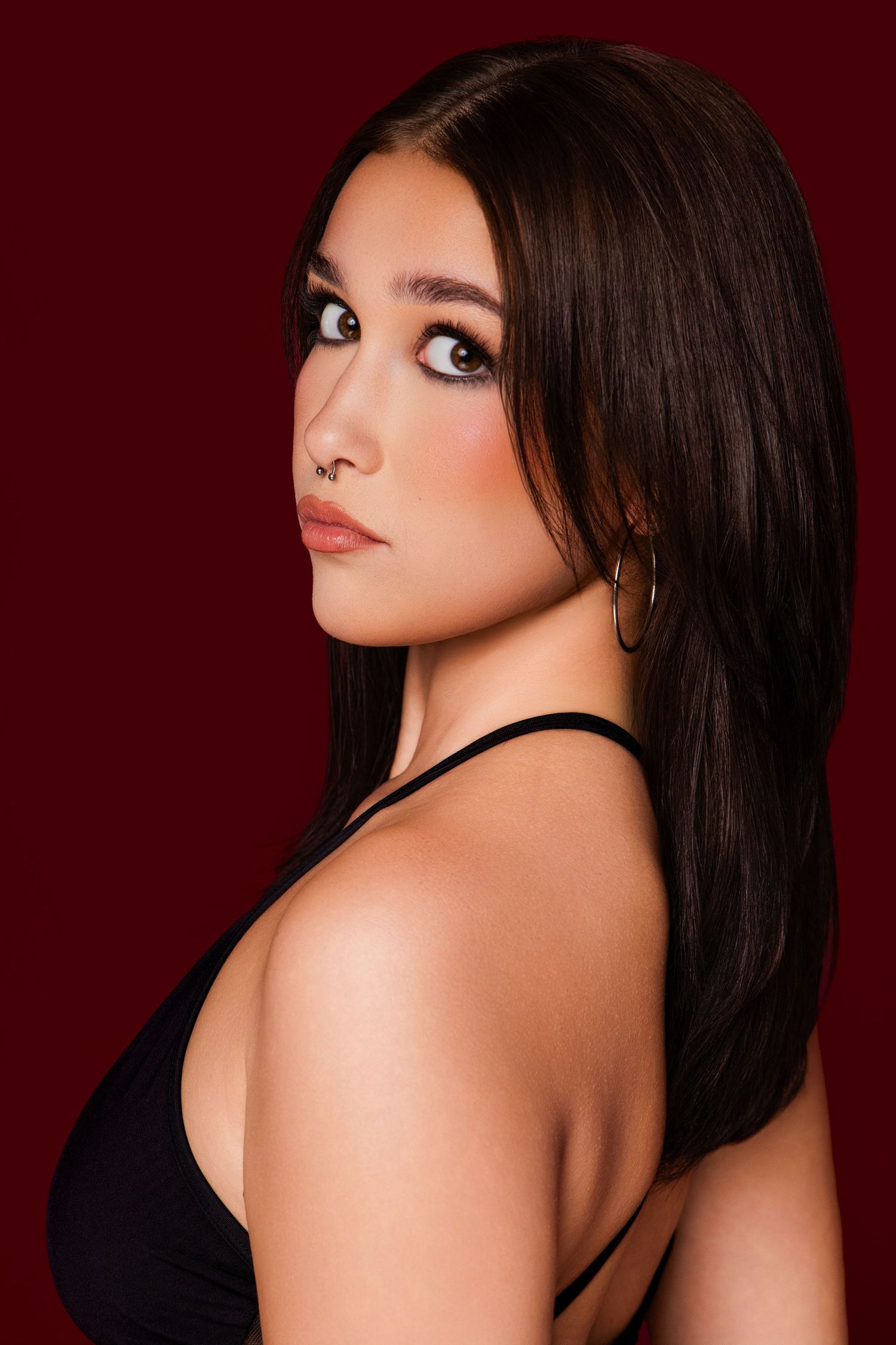 Young woman with long dark hair poses in black top against deep red background, wearing gold hoop earrings.