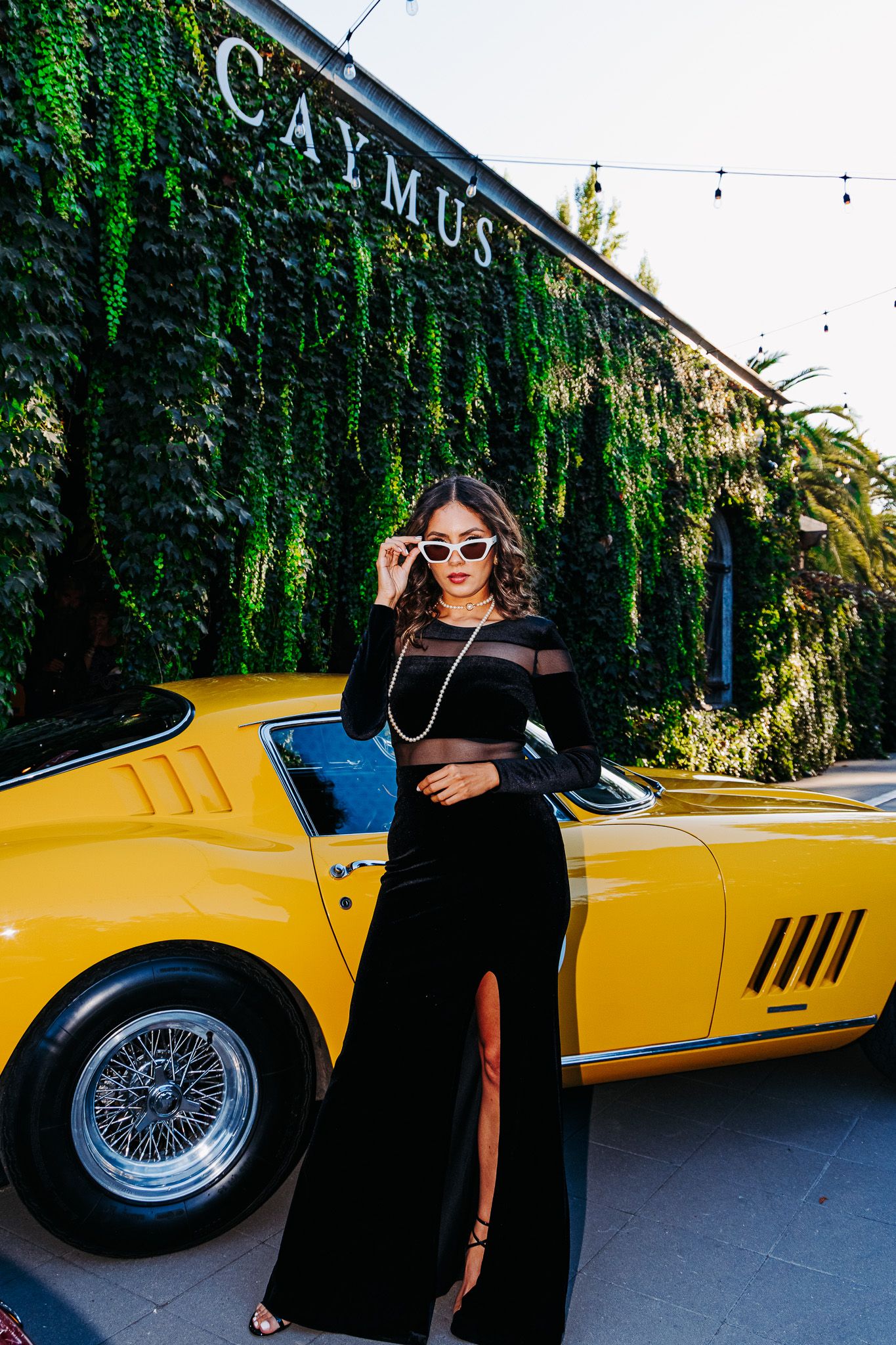 A series of four fashion photos featuring a black dress against a yellow vintage sports car with ivy-covered walls.