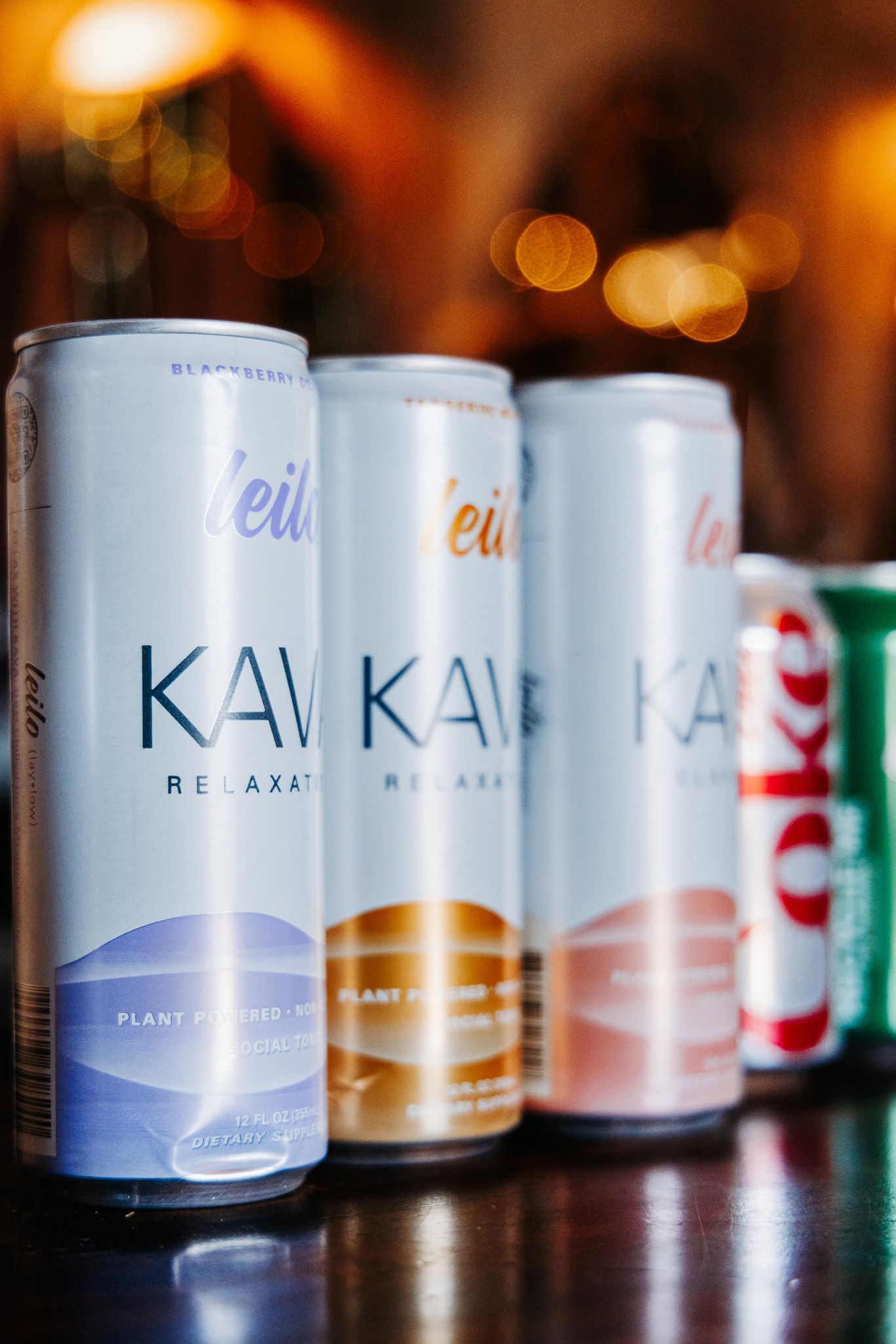 Row of Leilo Kava Relaxation drink cans displayed on a bar surface with bokeh lights in the background.