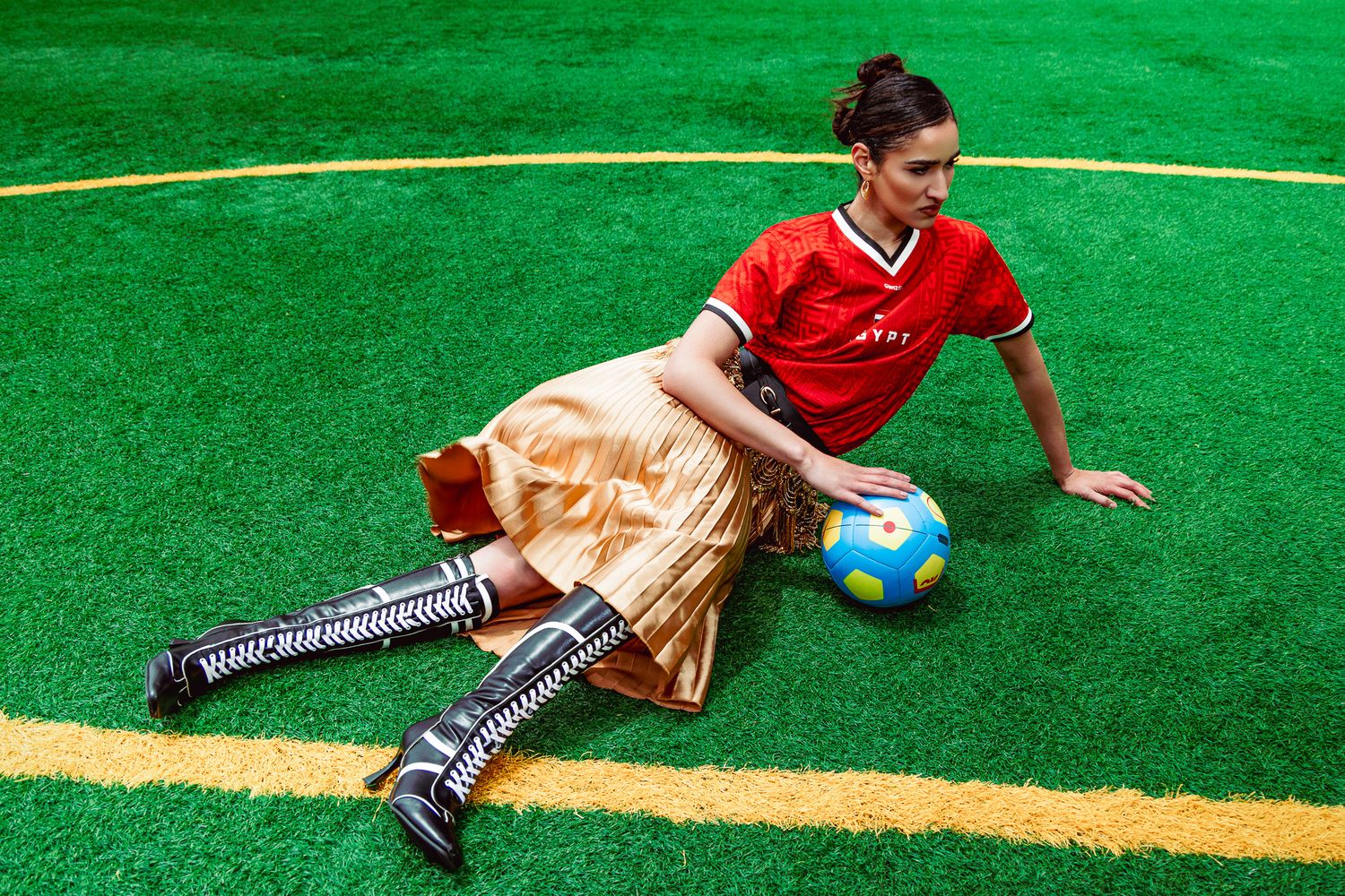 Woman in red soccer jersey, gold skirt, and tall boots sitting on green turf field with a soccer ball.