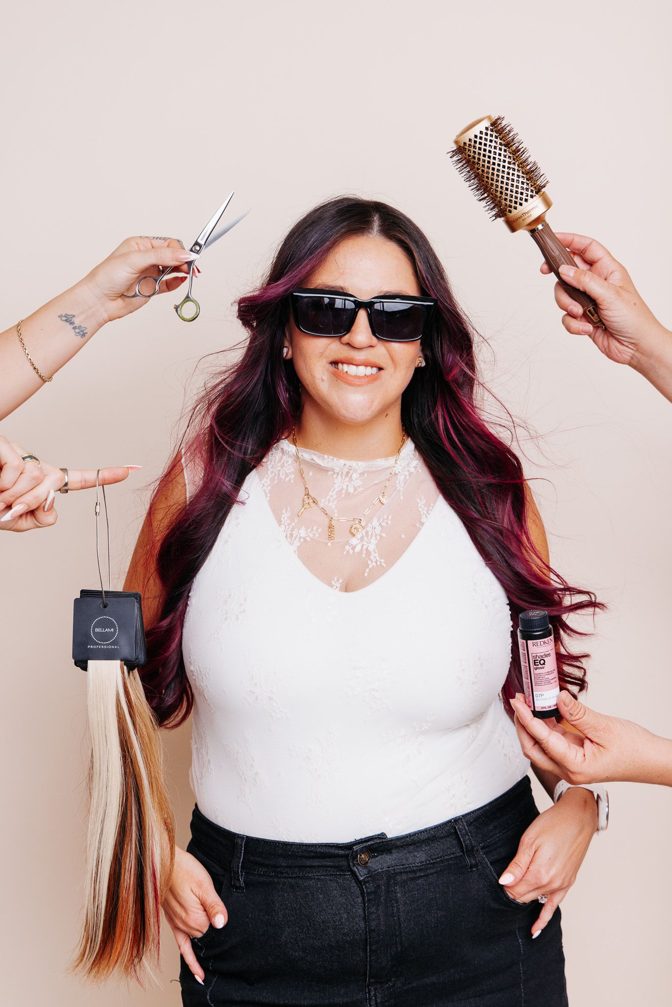 Woman with long pink-highlighted hair wearing sunglasses surrounded by hands holding hair tools and extensions.