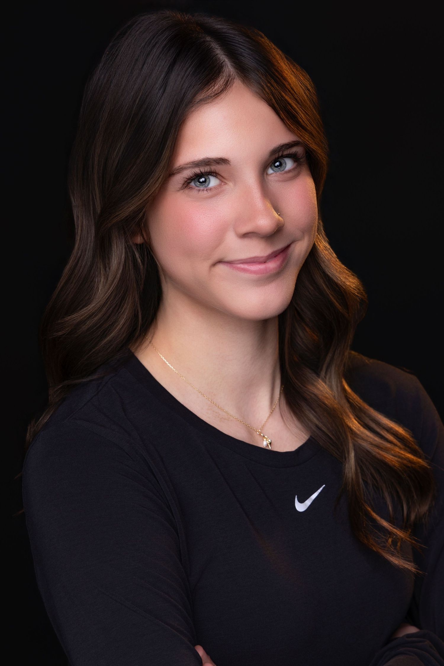 Professional portrait against dark background showing confident pose in black Nike athletic top with wavy brown hair.