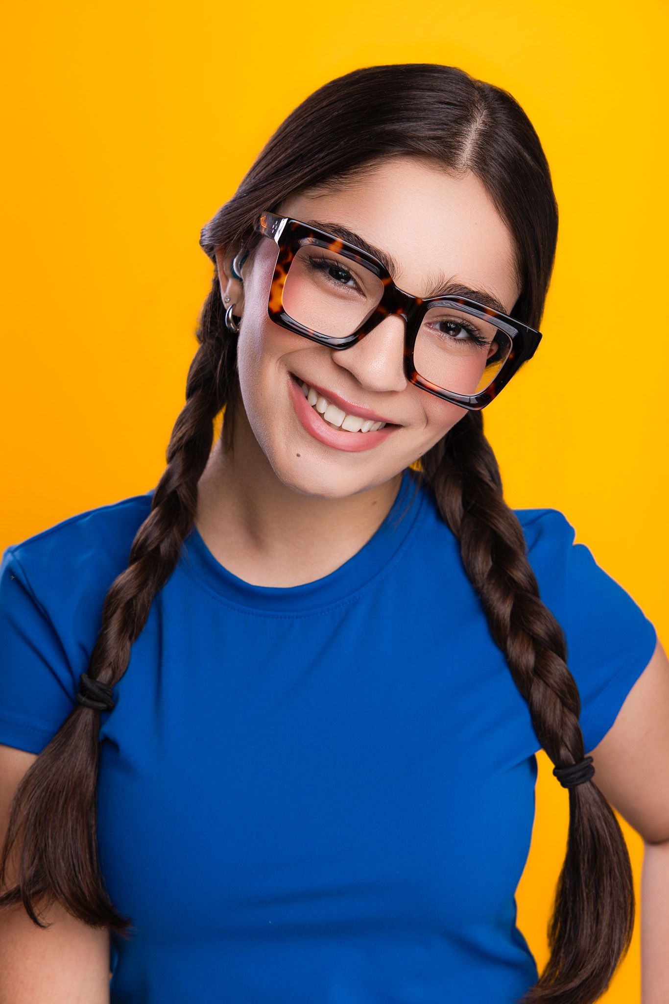 Smiling young woman with braids and large glasses wearing a blue t-shirt against a yellow background.