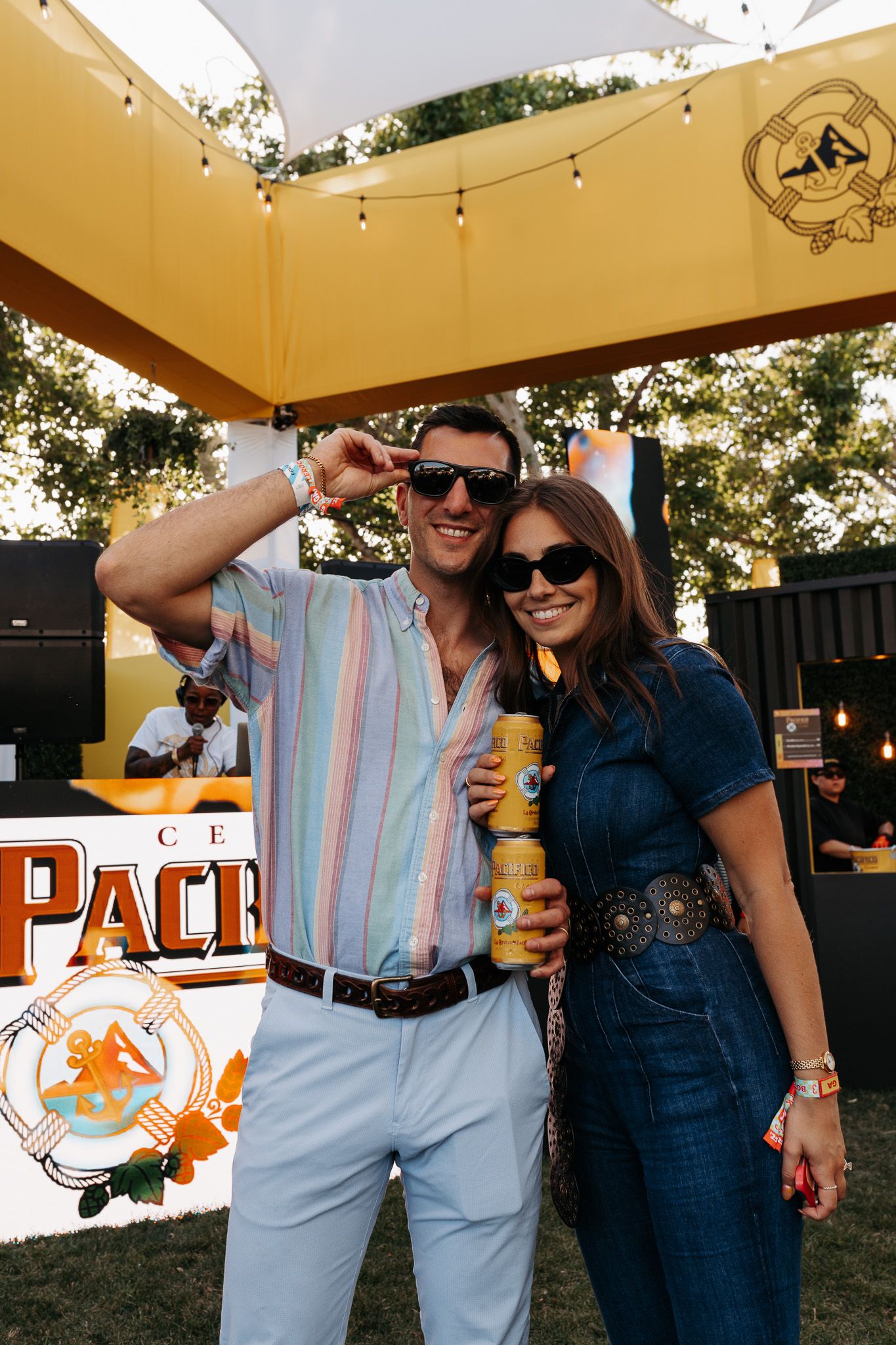 A couple enjoying drinks and smiling together at an outdoor event with string lights and signage in the background.
