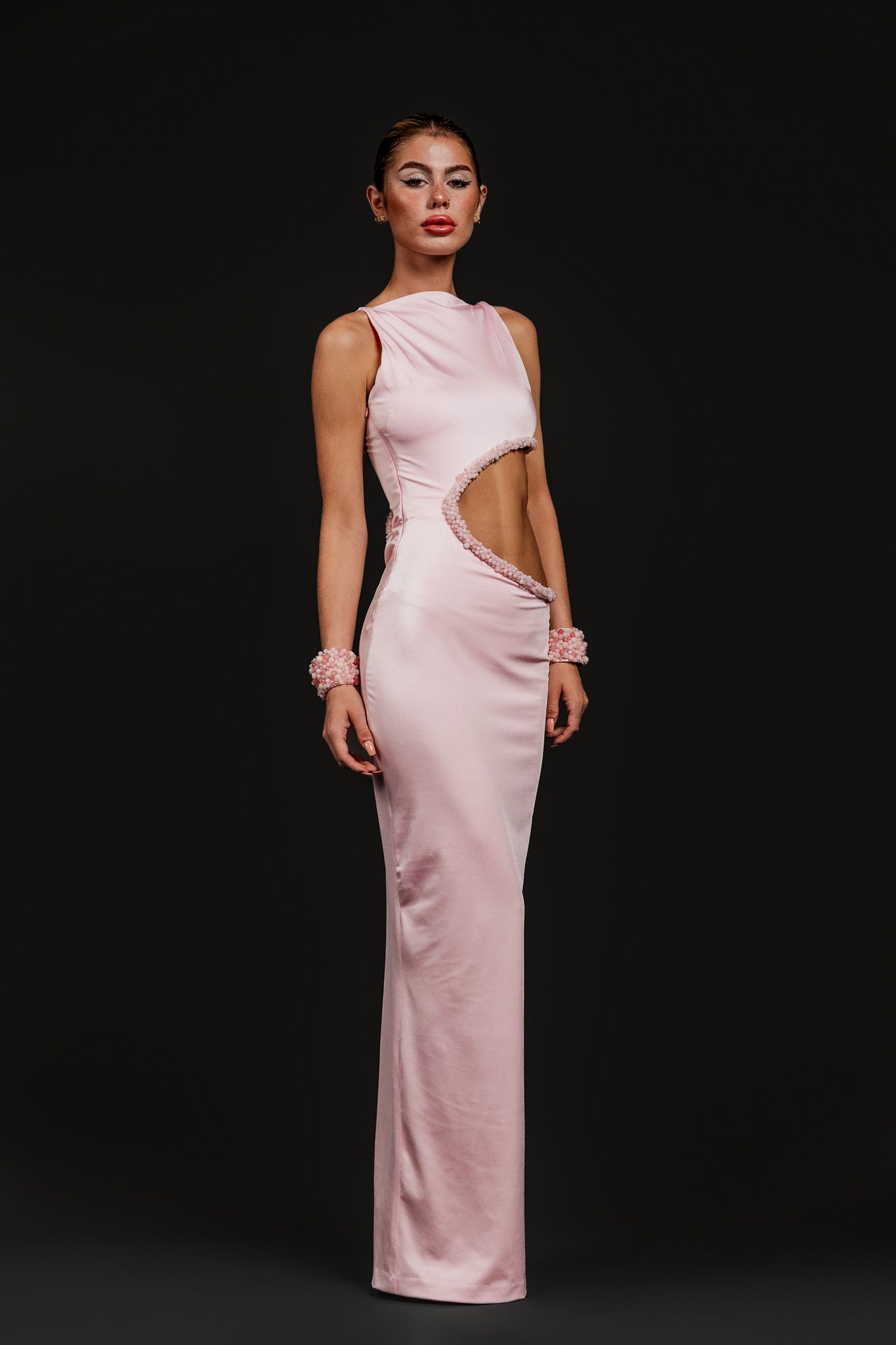 Woman in sleeveless pink satin gown with cutout waist detail and floral wrist cuff on dark background.