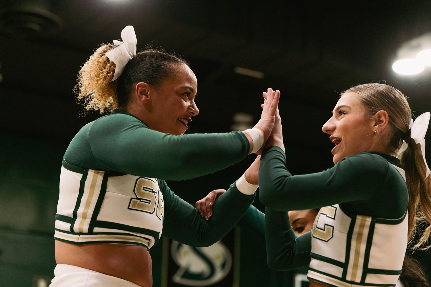 Two cheerleaders in green uniforms high-fiving and smiling during a cheerleading event.
