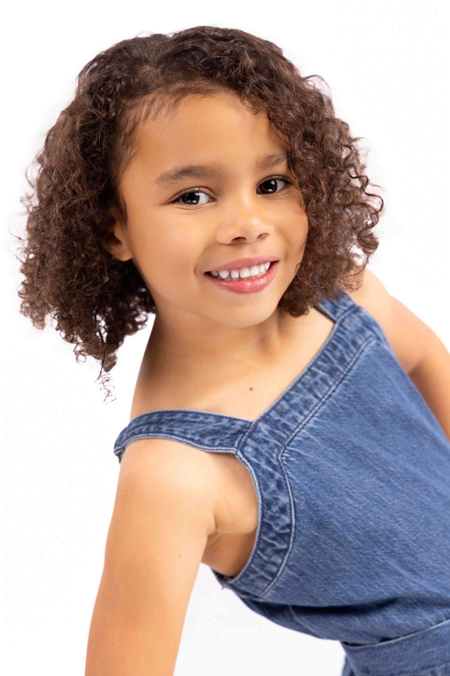Child model with curly hair poses playfully in denim outfit against white background.
