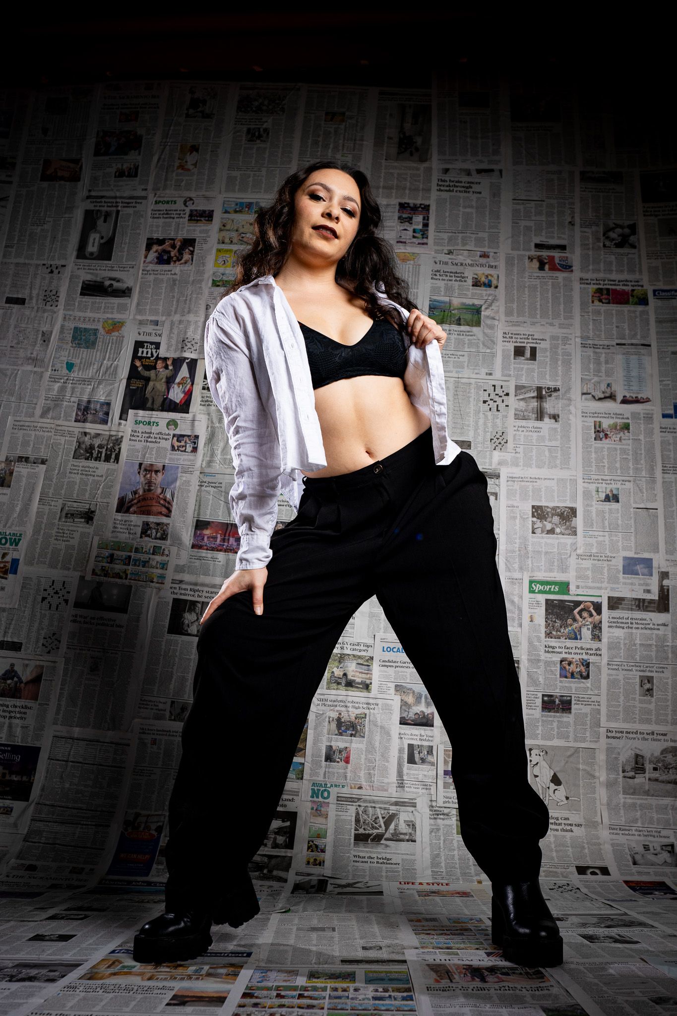 Dancer in black cargo pants and white jacket poses confidently against newspaper-covered wall in dramatic lighting.