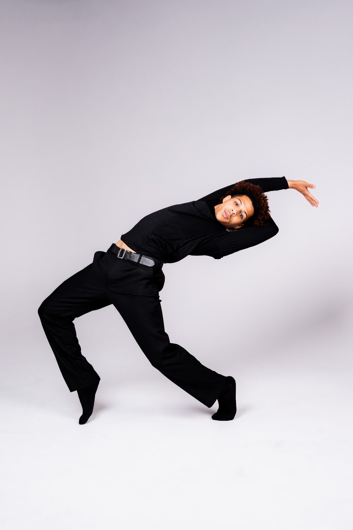 A dancer in black clothing performs a dynamic backward bend against a gray studio background.