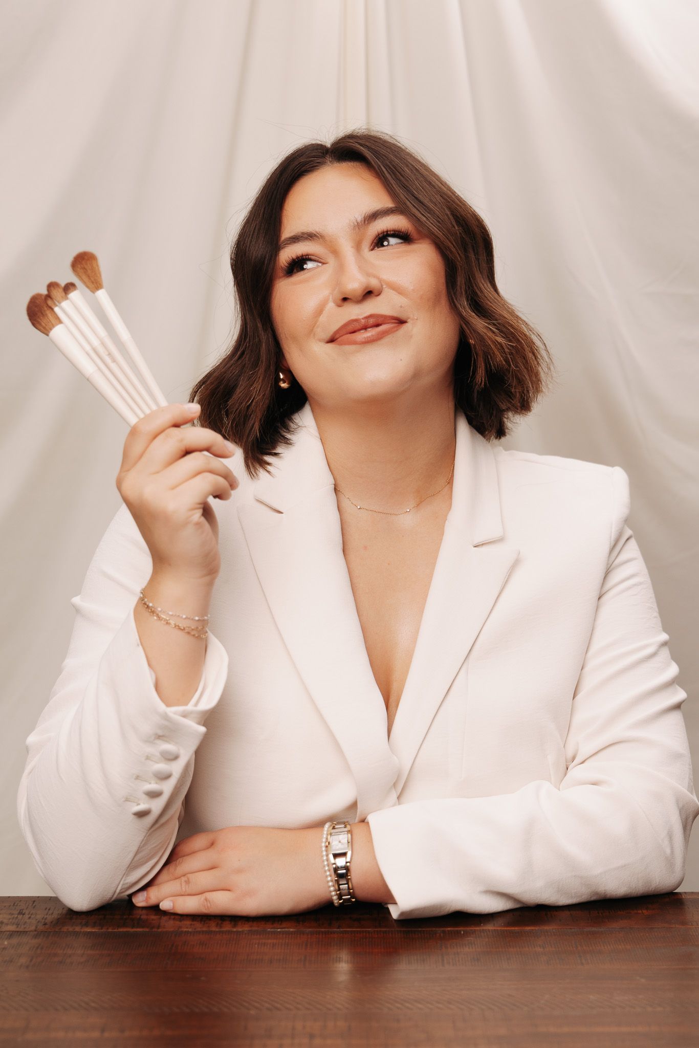 Professional portrait series showing someone in a white blazer laying on wooden surface holding makeup brushes in different poses.