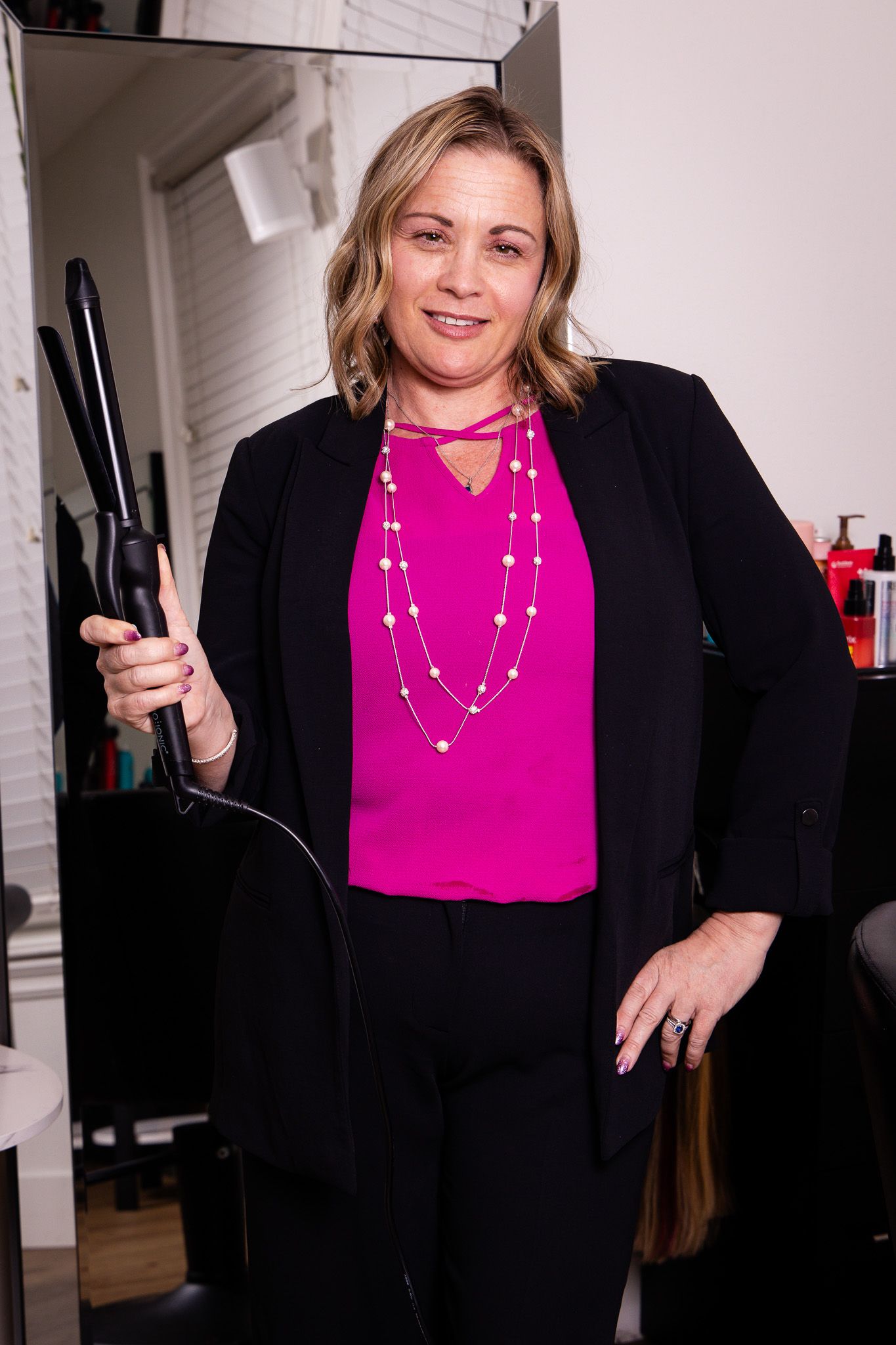 Hair stylist in black blazer and bright pink top with layered necklace holds styling tool in salon.