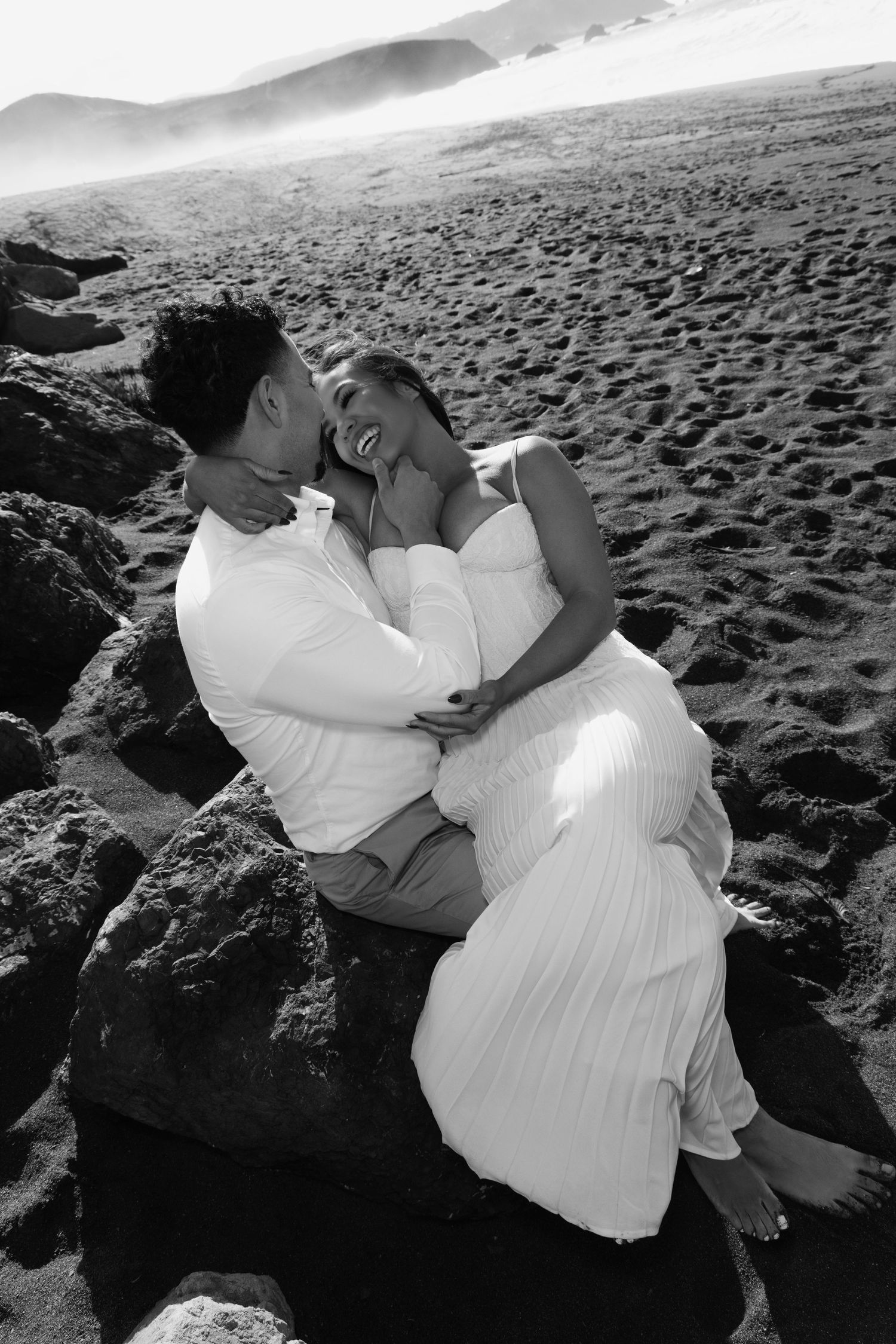 A romantic couple in white attire embraces on dark rocks by the beach at sunset in a series of black and white photos.