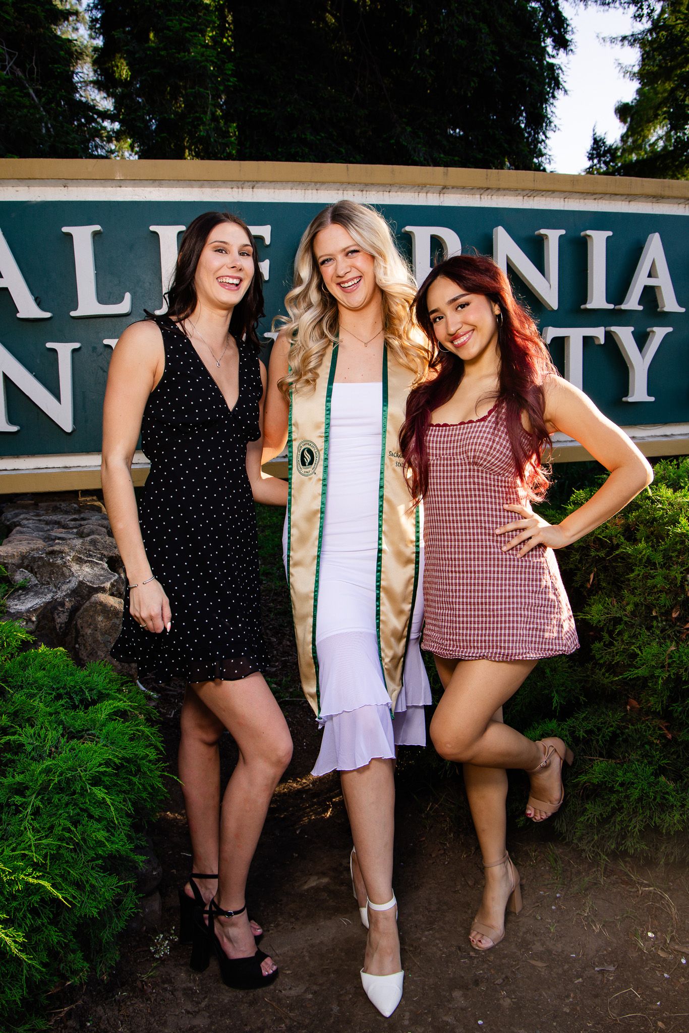 Three young women posing in front of a California University sign, one wearing a graduation stole.