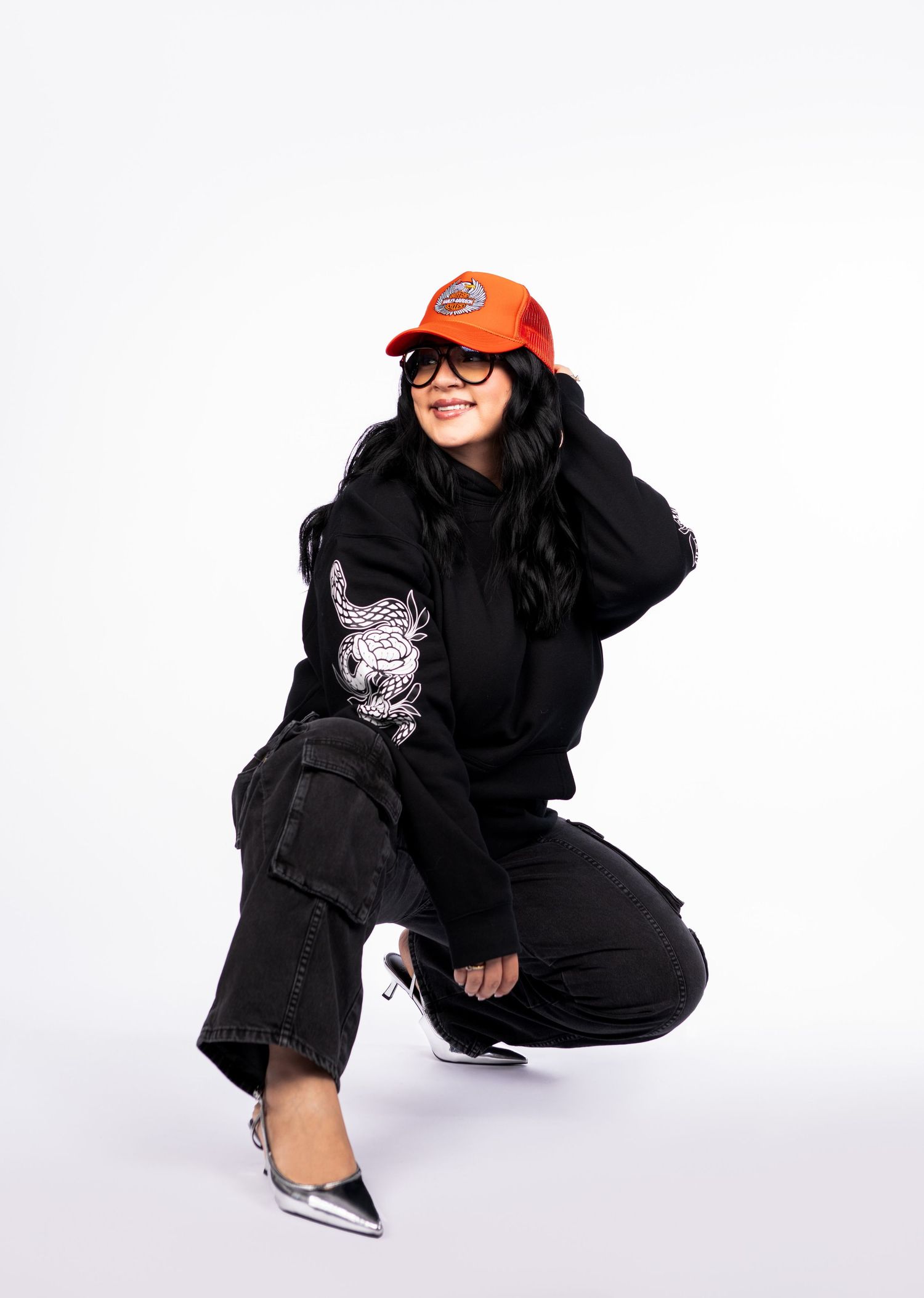 Woman crouching in black graphic hoodie and jeans, wearing orange cap and silver heels on white background.