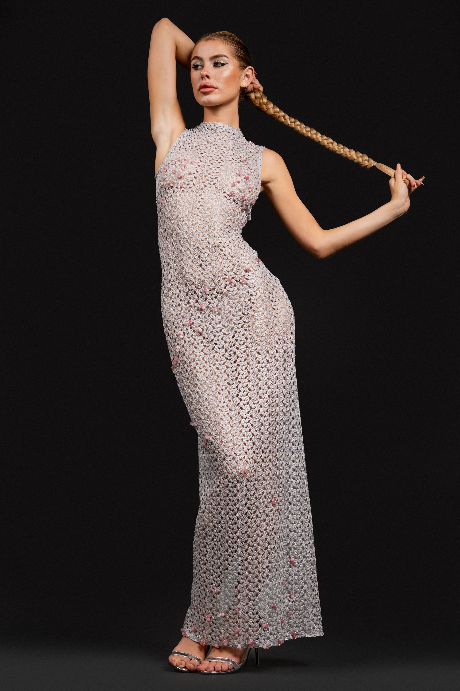 Model in sheer white floral maxi dress with mock neck, posing with long braid and one arm raised against dark background.