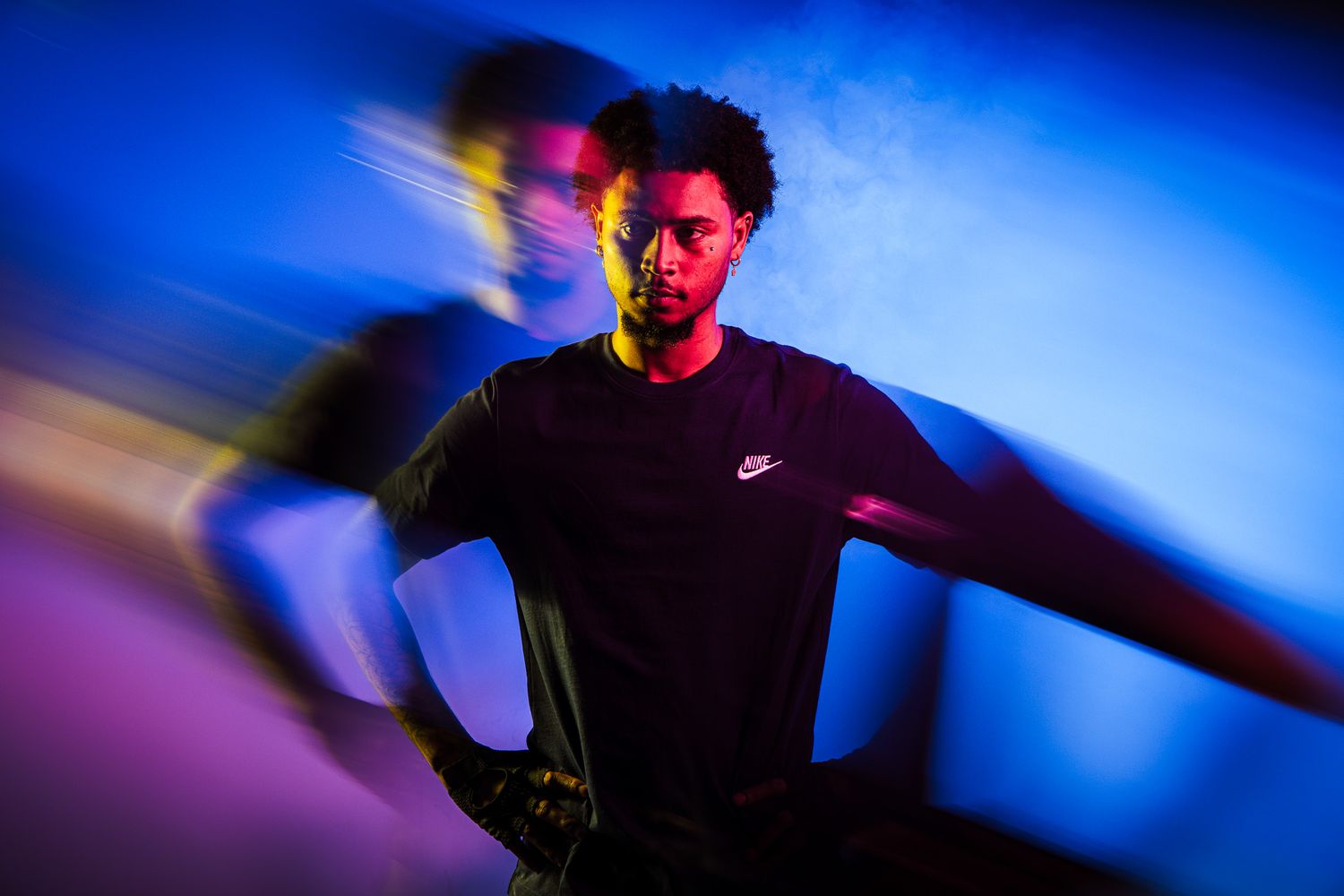 Man in black Nike shirt poses dramatically under vibrant blue and pink studio lighting with motion blur effect.