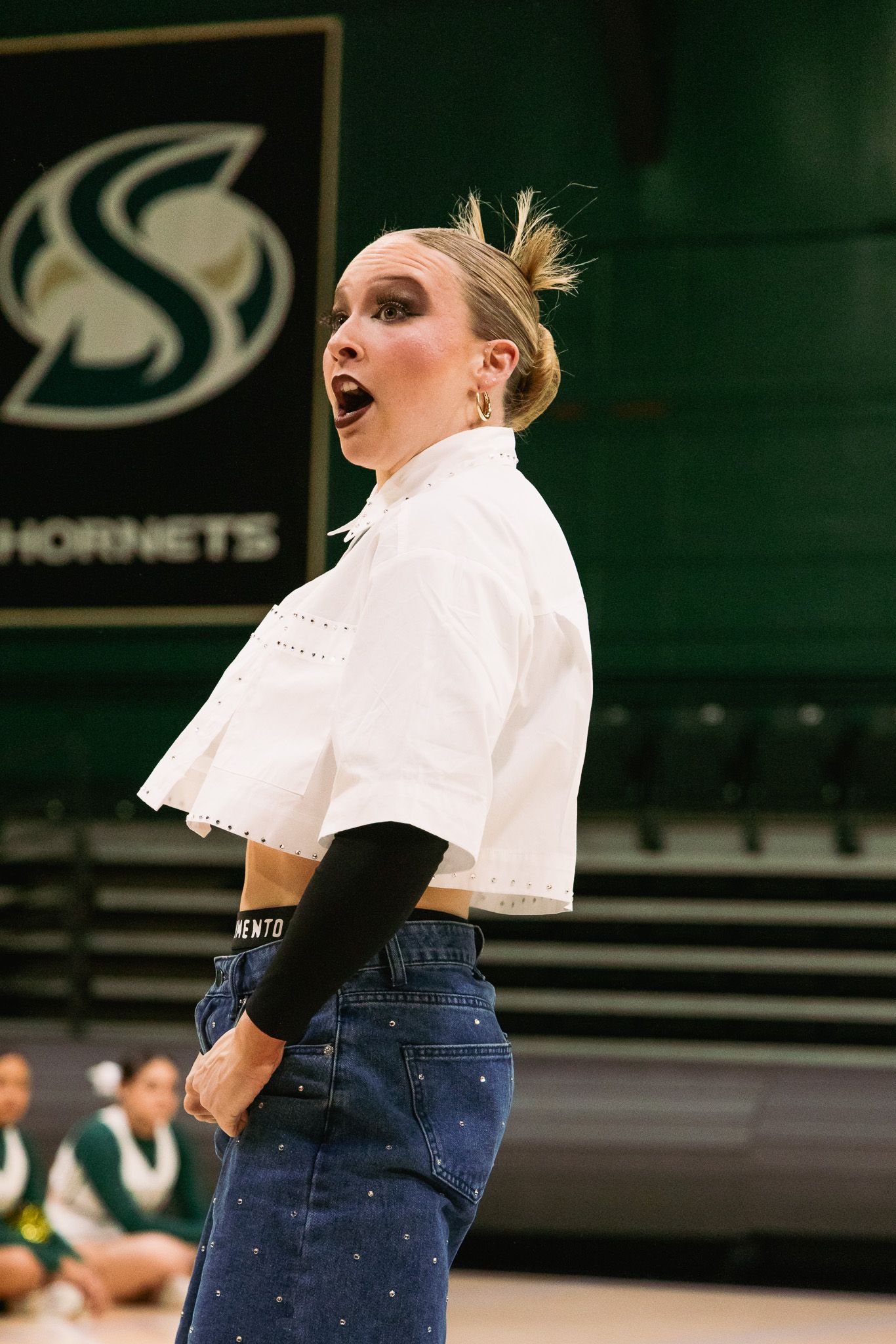 Young woman in white crop top and jeans performing at Sacramento State Hornets arena with expressive face.