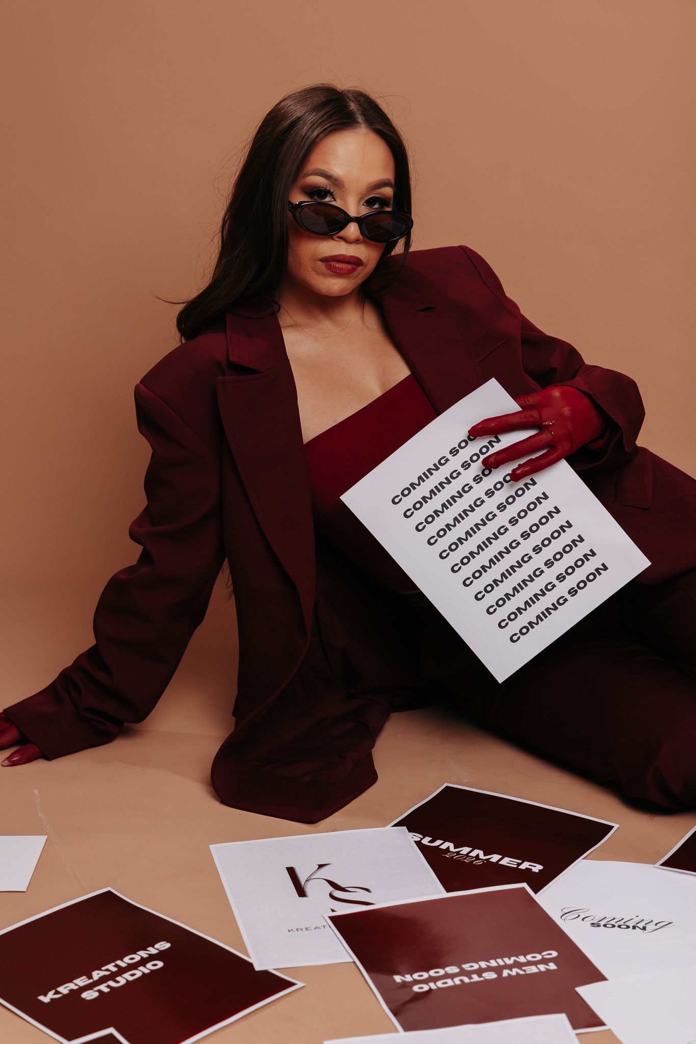 Stylish woman in burgundy suit and sunglasses poses on floor with Kreations Studio coming soon promotional papers.