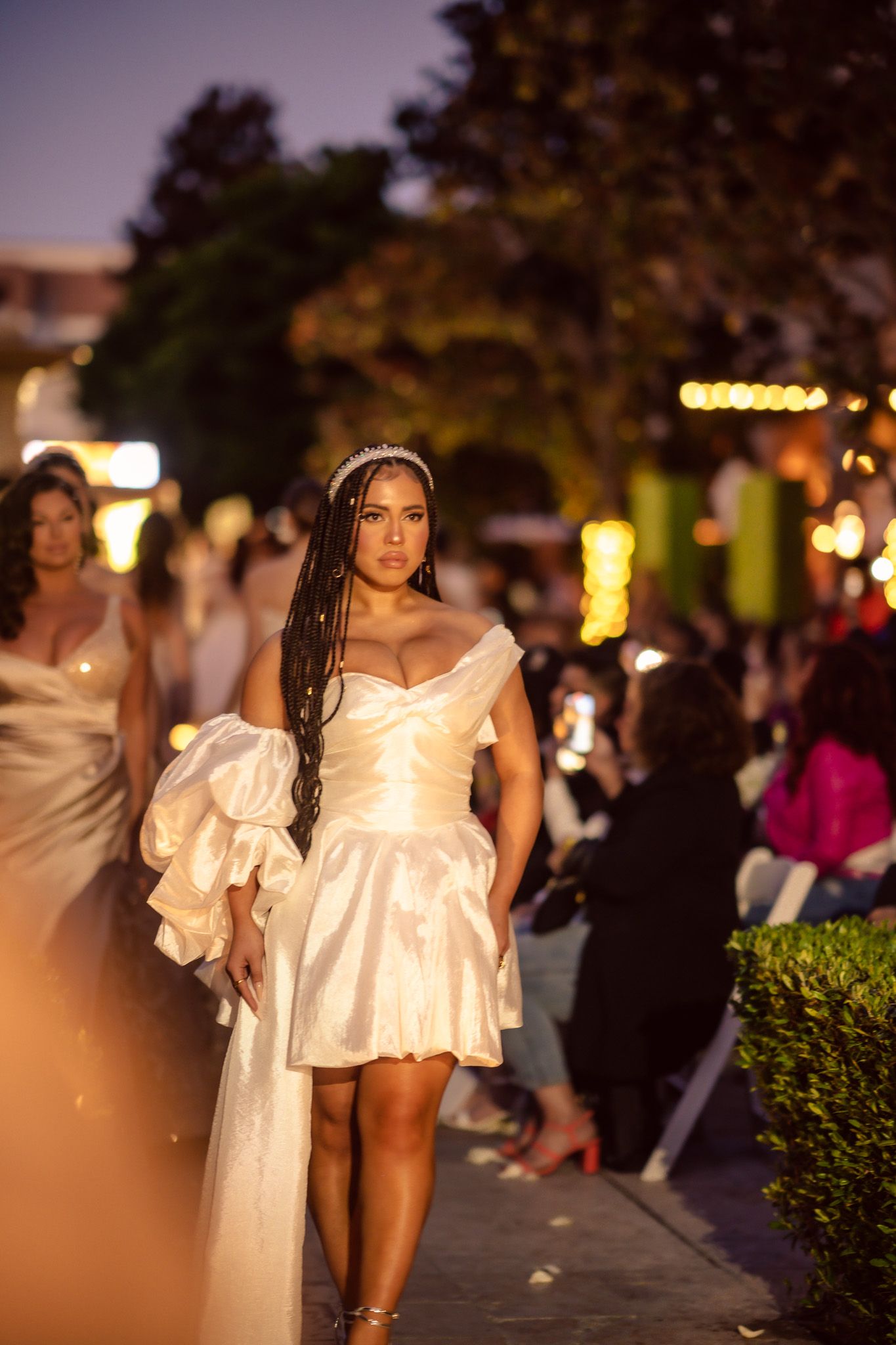 Model presents short white off-shoulder dress with dramatic train at evening outdoor fashion show.