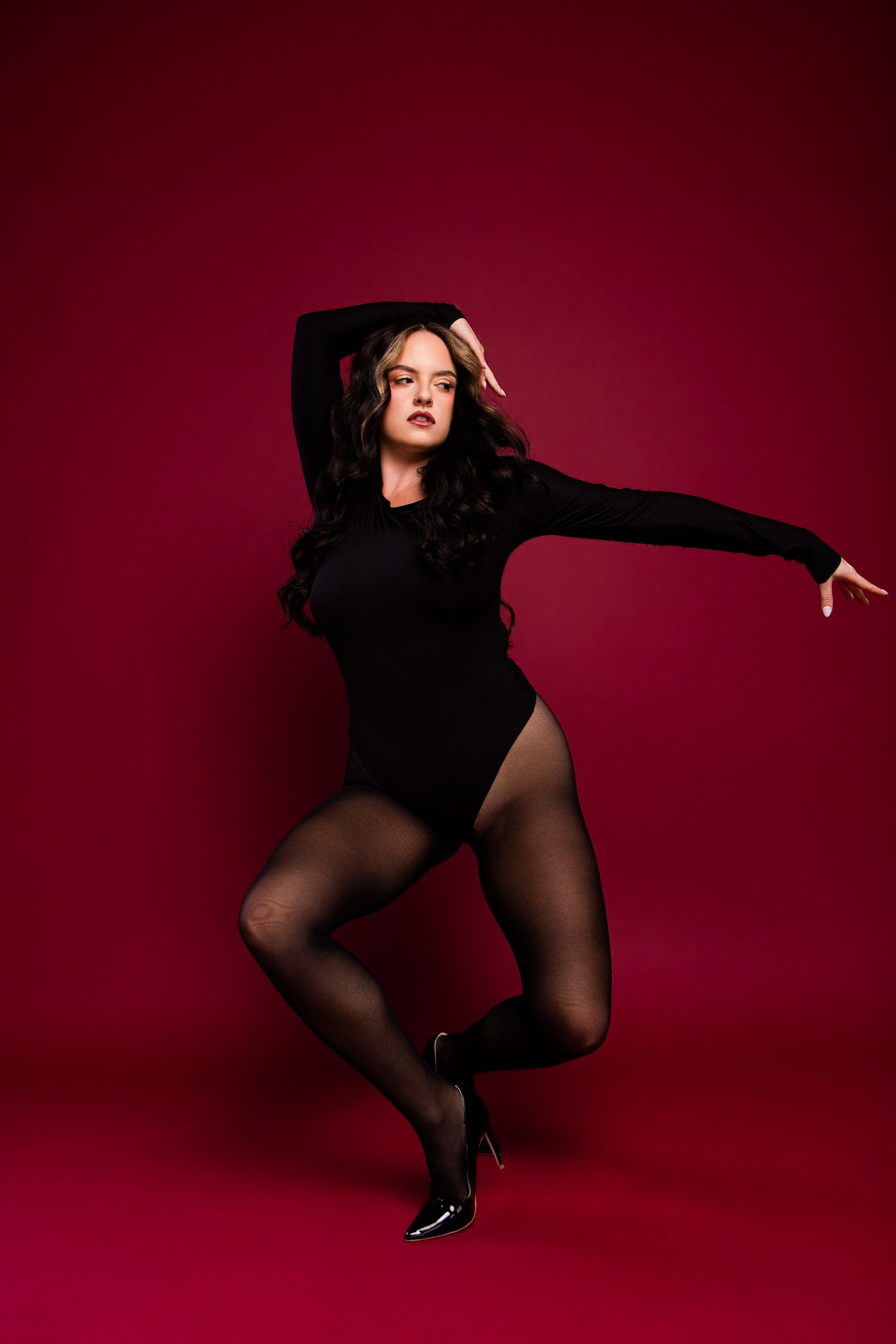 Woman in black bodysuit and tights poses dynamically against a deep red background in high heels.