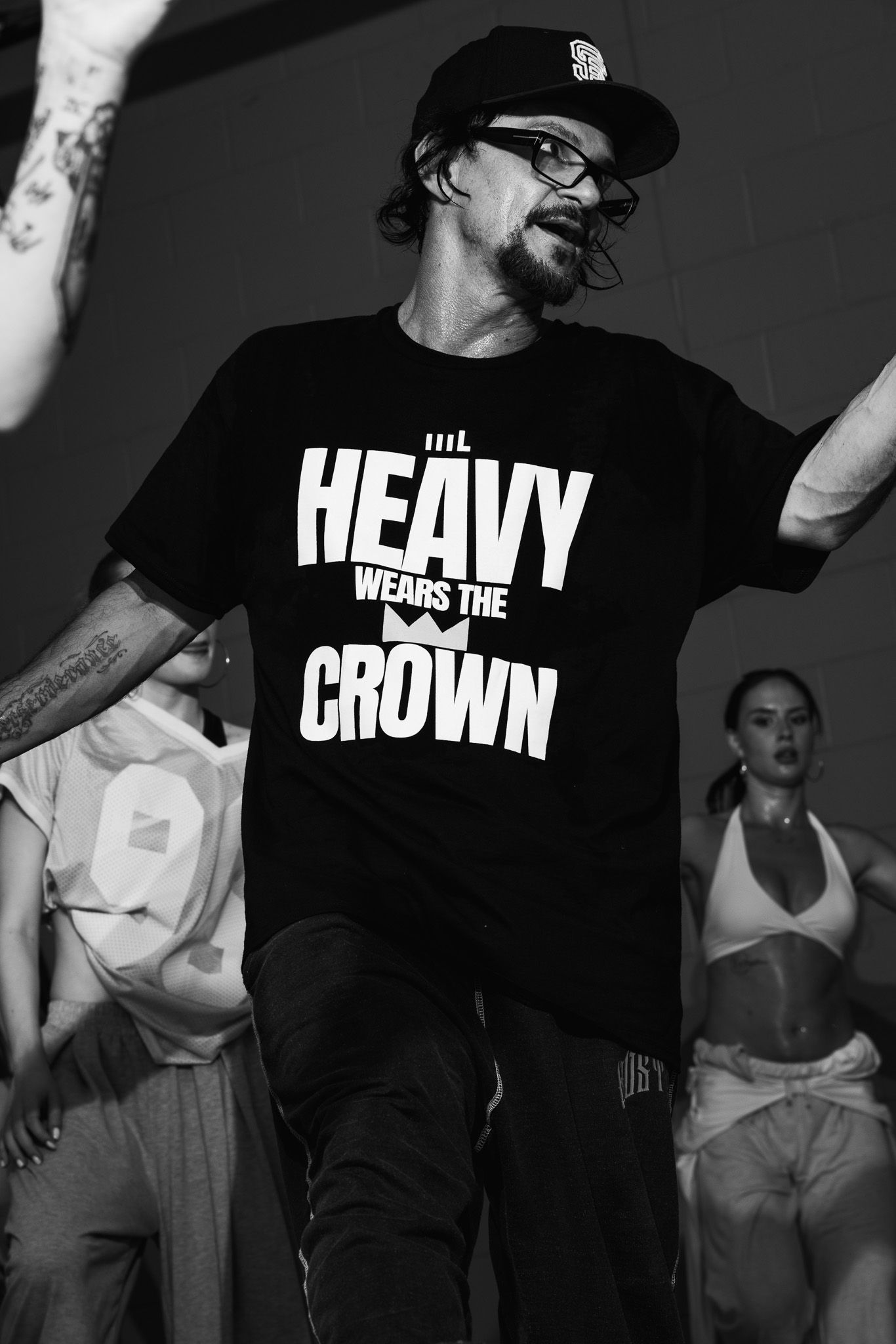 Man in black Heavy Wears The Crown t-shirt and cap dancing on stage in black and white photo.