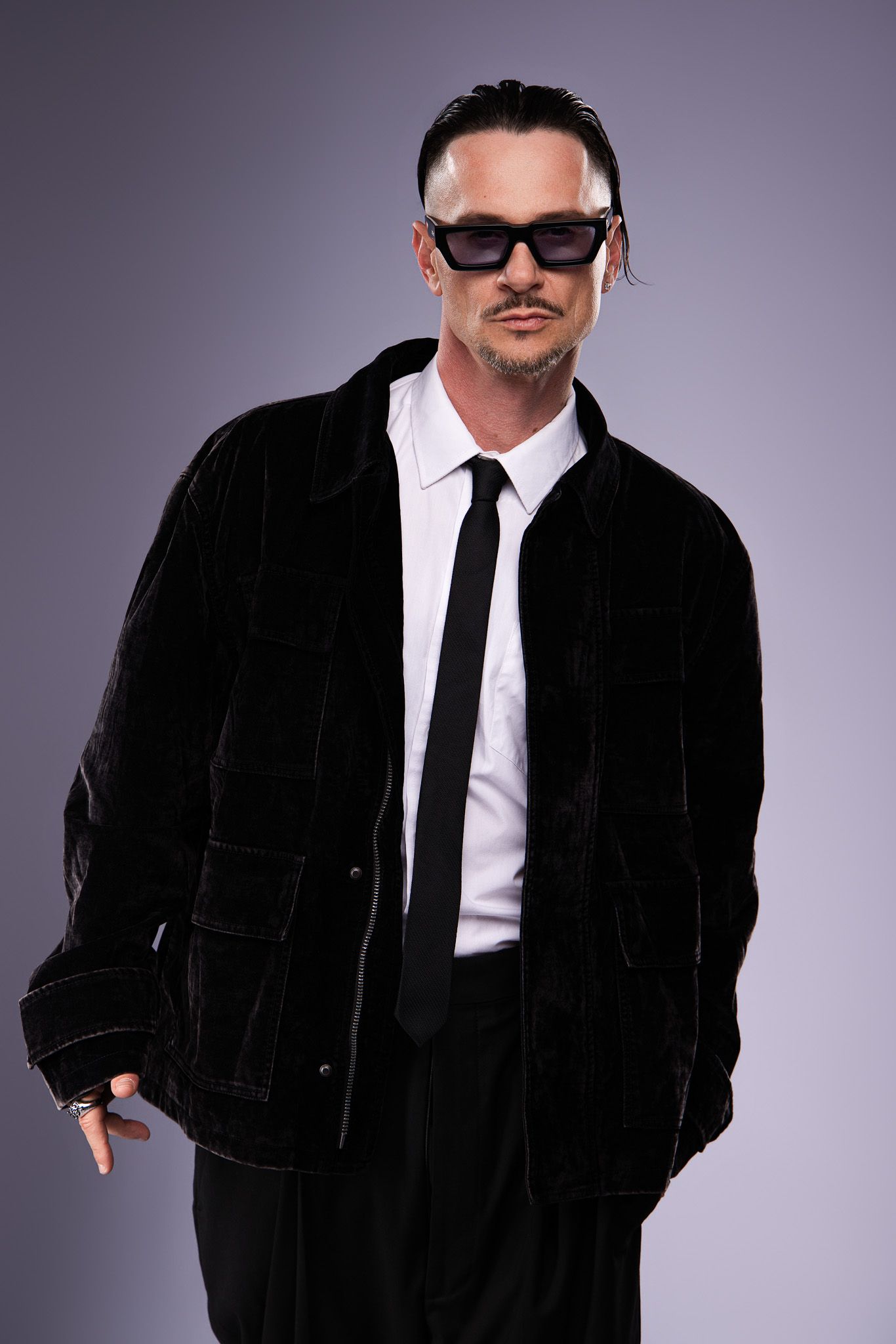 Stylish man in black velvet jacket, white shirt, tie, and sunglasses posing confidently against gray background.