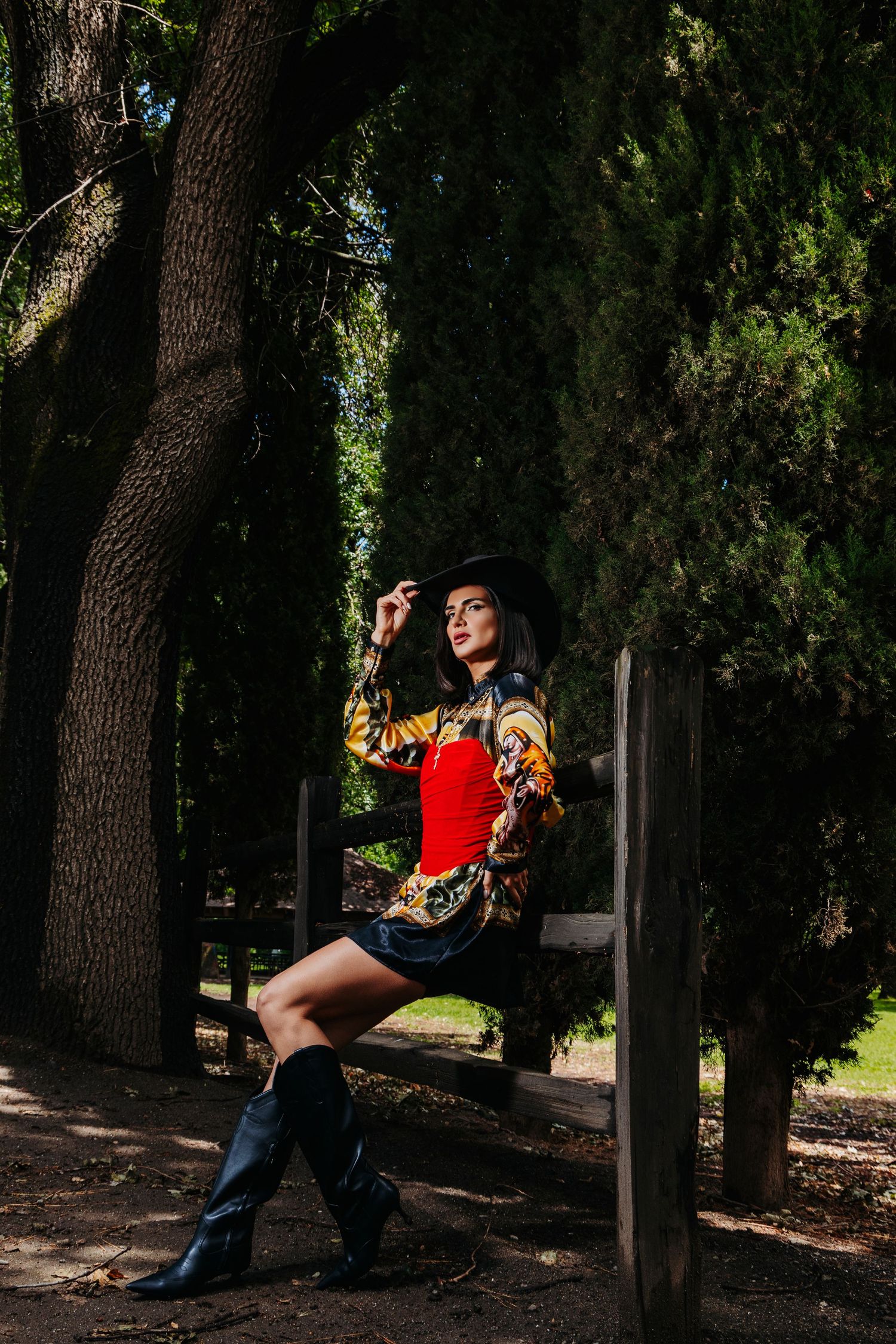 Woman in black hat, red corset, patterned jacket, and knee-high boots poses on wooden fence in shaded park.