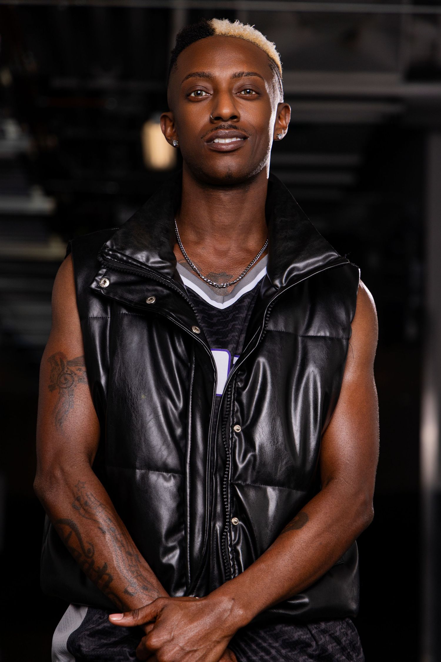 Young Black man with blonde-tipped hair wearing a black leather sleeveless vest and silver chain necklace, arms crossed.