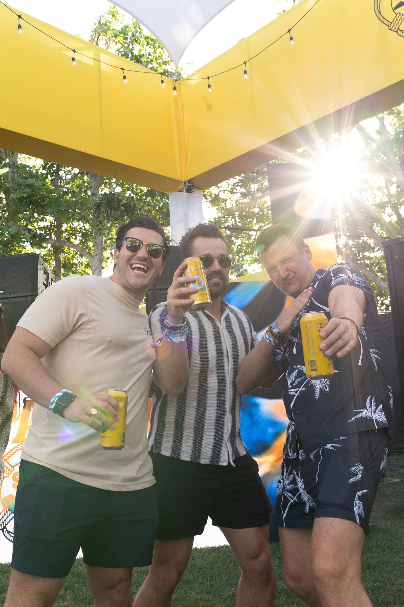 Three men smiling and holding yellow drinks at an outdoor festival under a sunny yellow canopy.