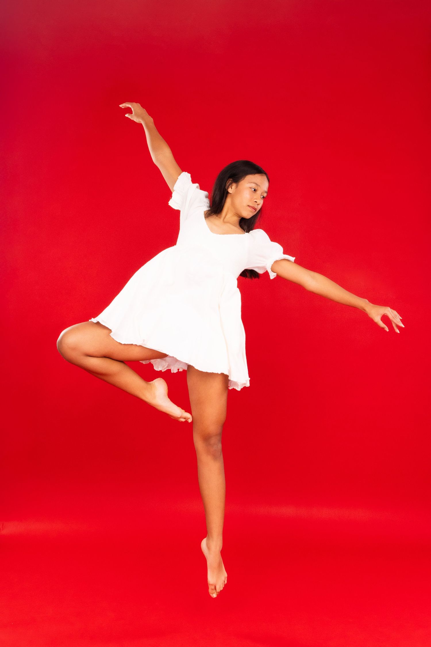 A dancer in a white dress performs graceful movements against a vibrant red studio backdrop.