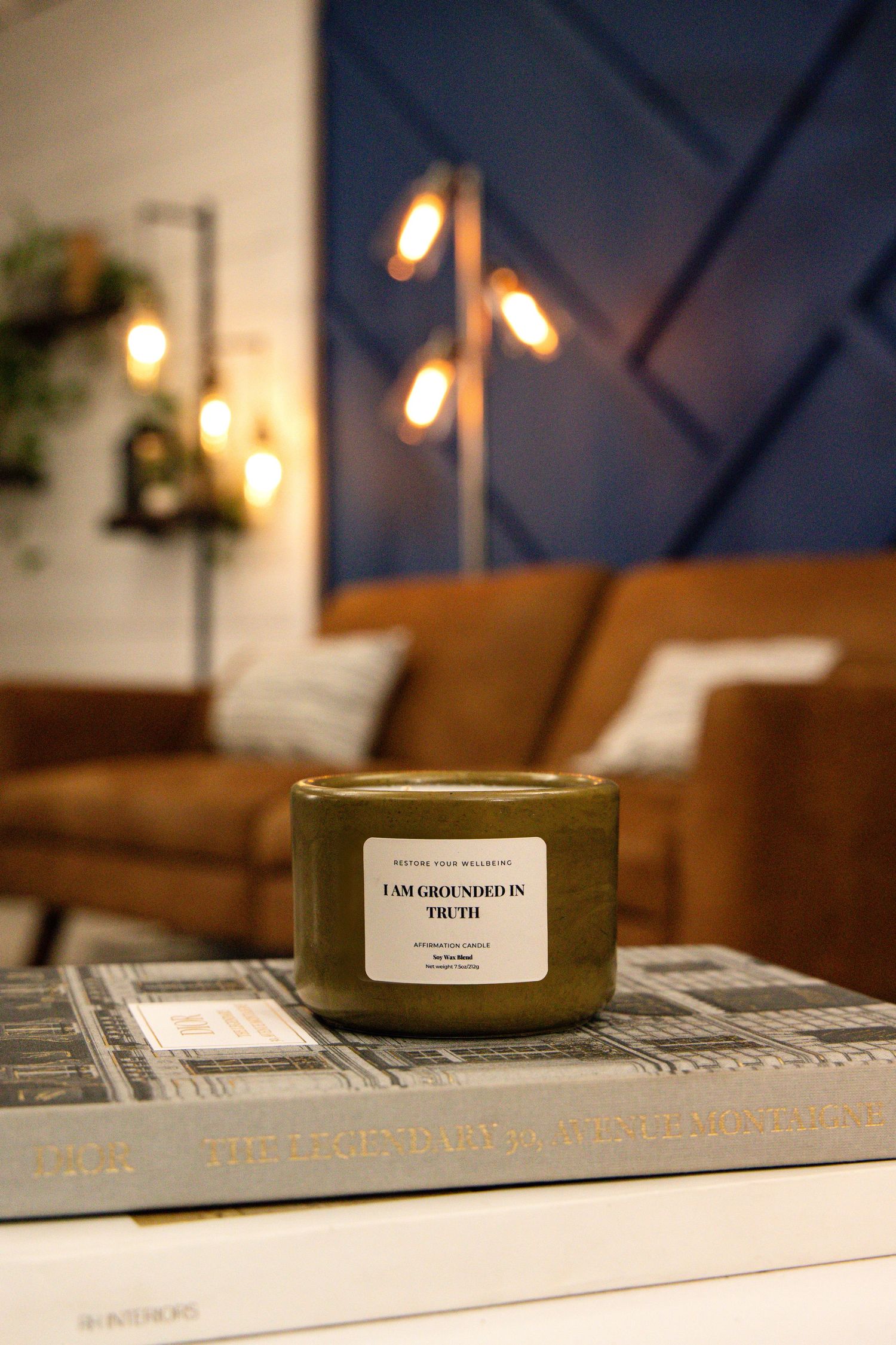A green scented candle sits on a coffee table book with warm lighting and a brown leather couch in the background.
