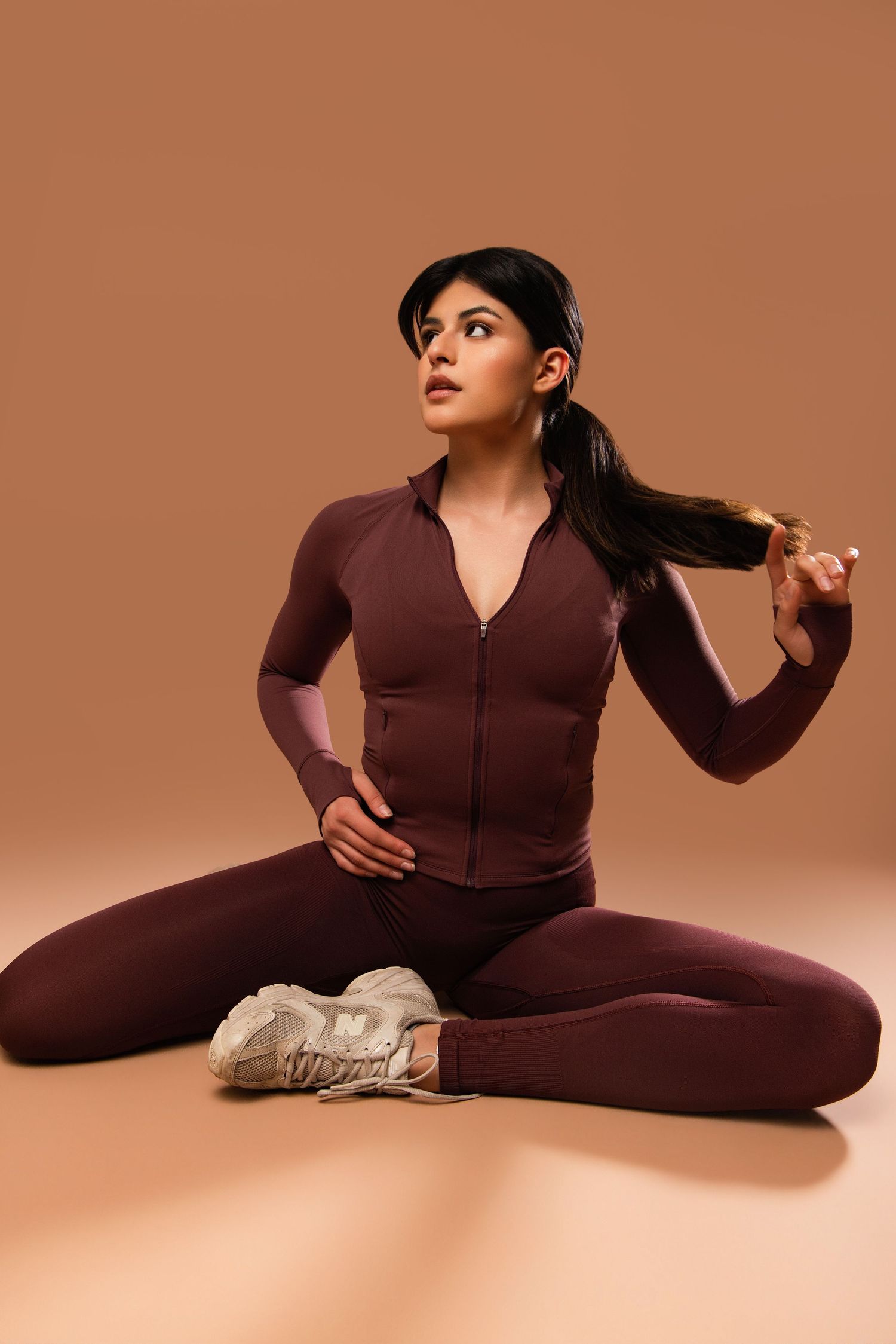 Athletic model in burgundy workout set with zip-front top and leggings poses on brown background with white sneakers.