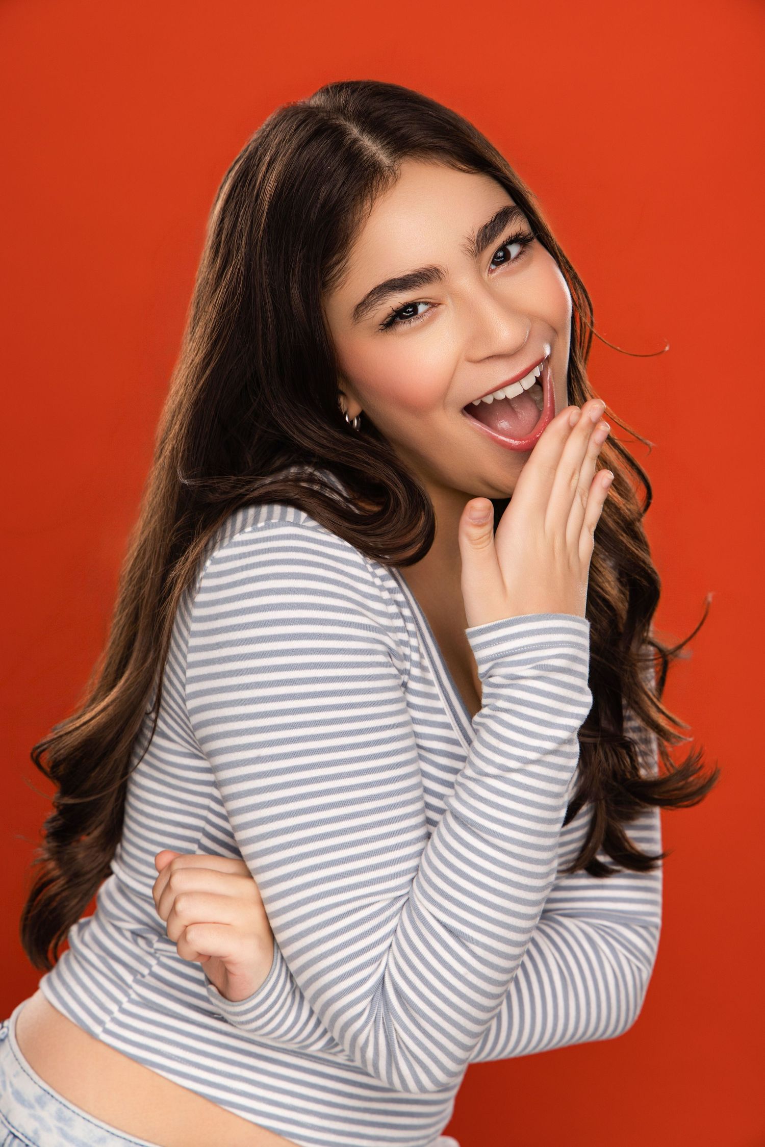 Fashion model poses playfully against vibrant orange background wearing striped top with flowing dark hair.