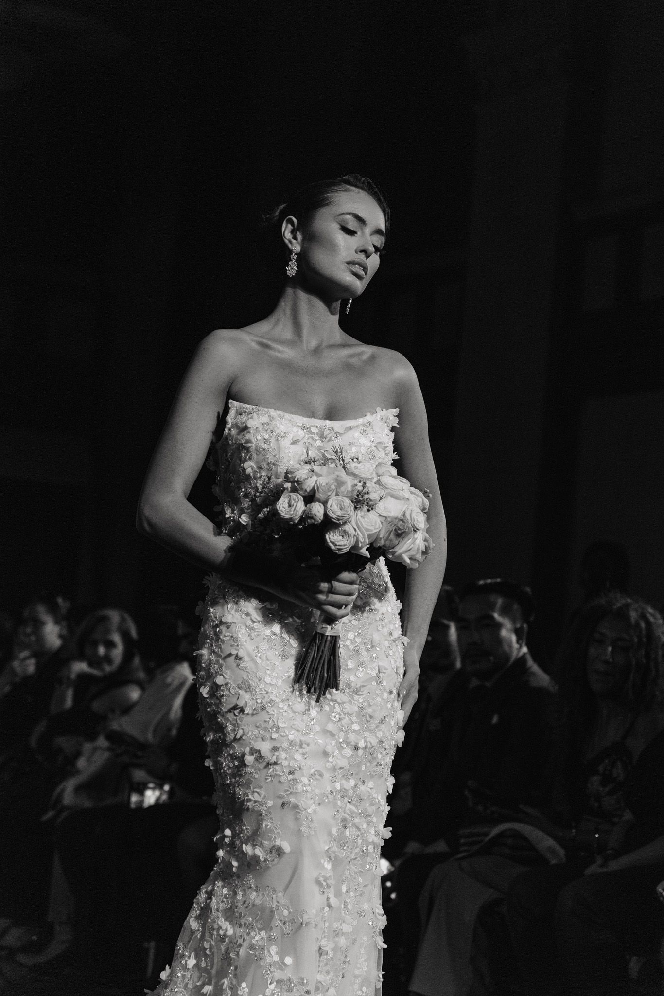 A model walks the runway in an elegant strapless wedding gown adorned with floral appliques and holding a bouquet.