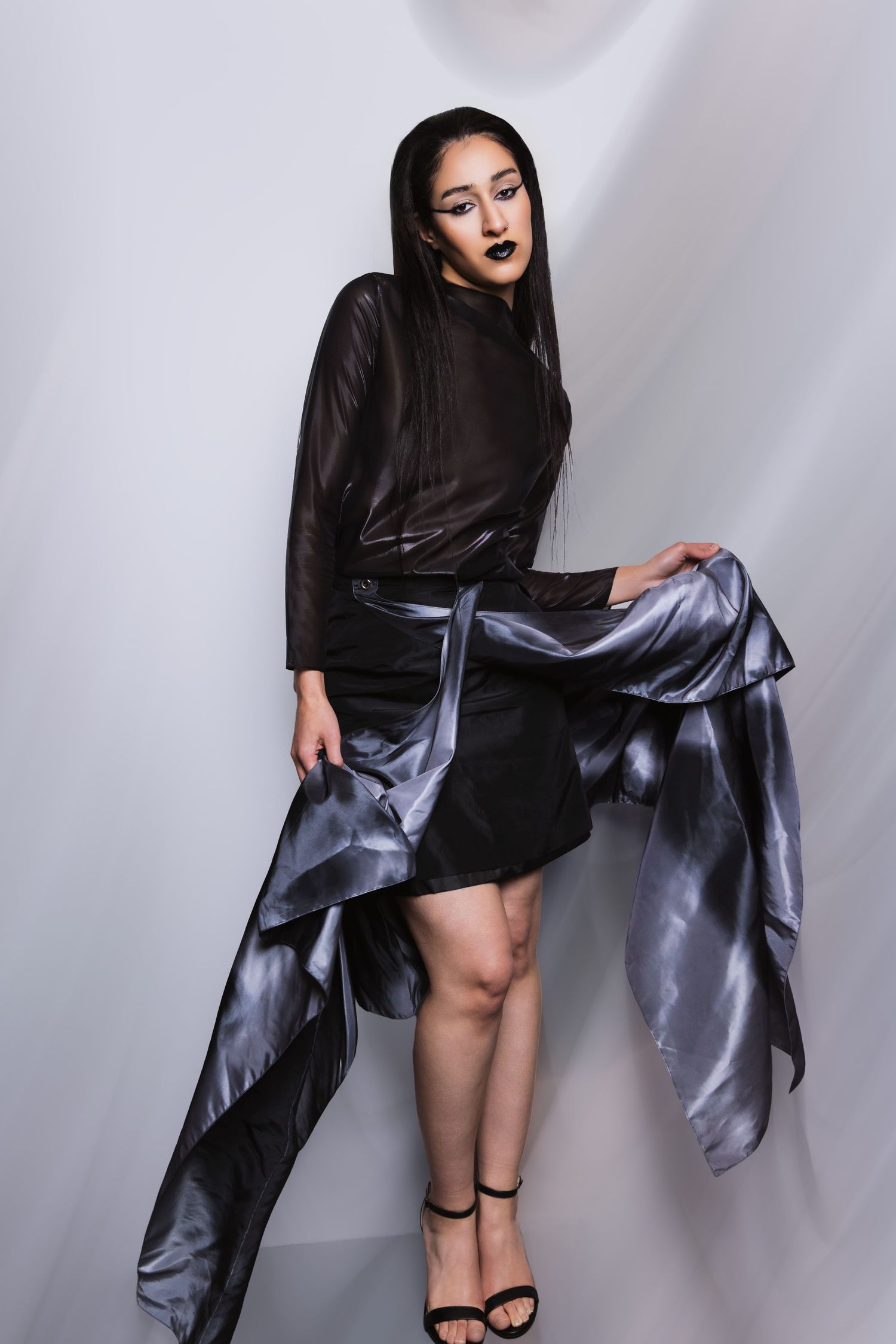 Fashion model in flowing black dress and dramatic fabric poses dynamically against white studio backdrop.